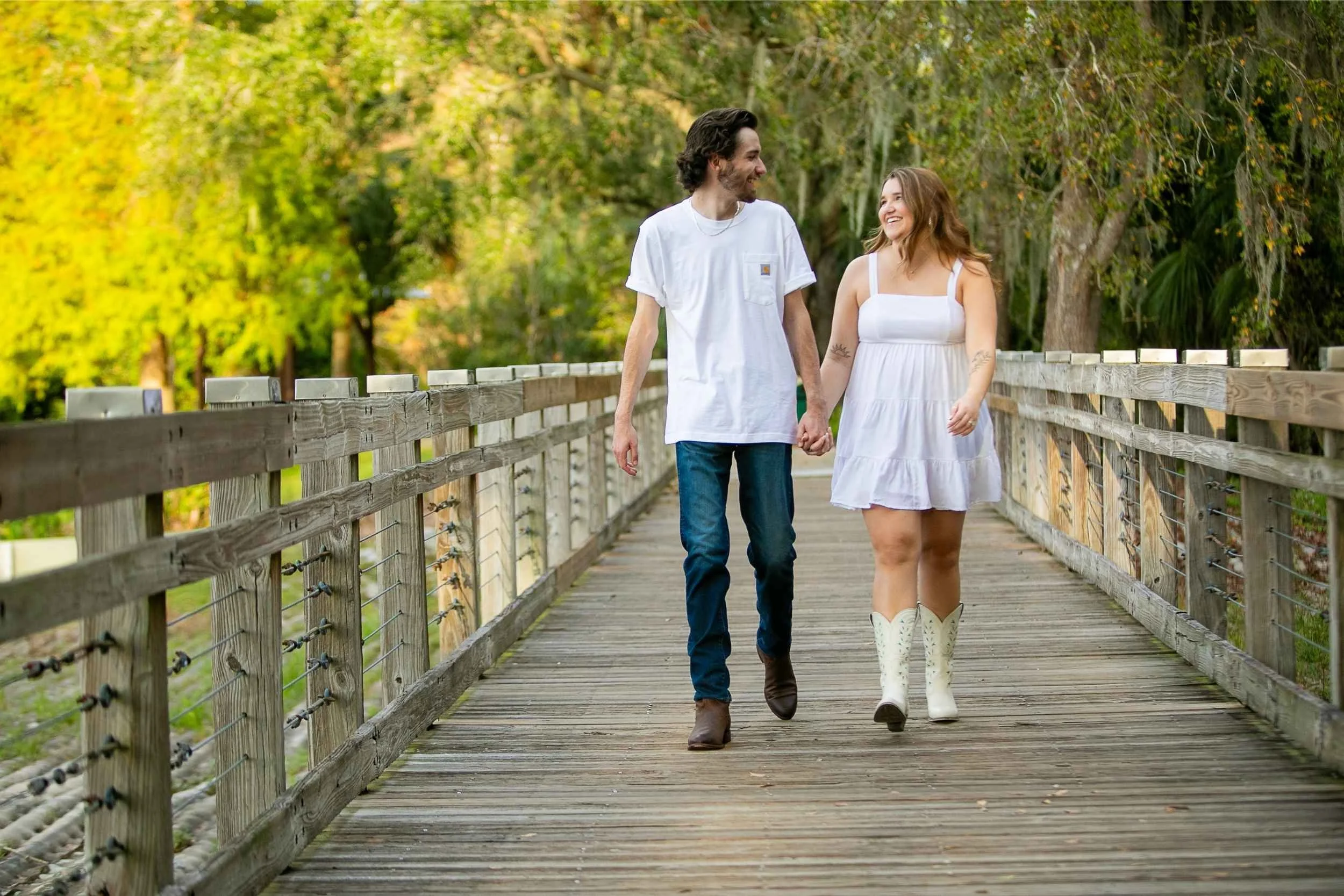 A couple holding hands and walking on a wooden bridge surrounded by trees in a park, smiling at each other for an engagement photography session with Cannonfire Photography.
