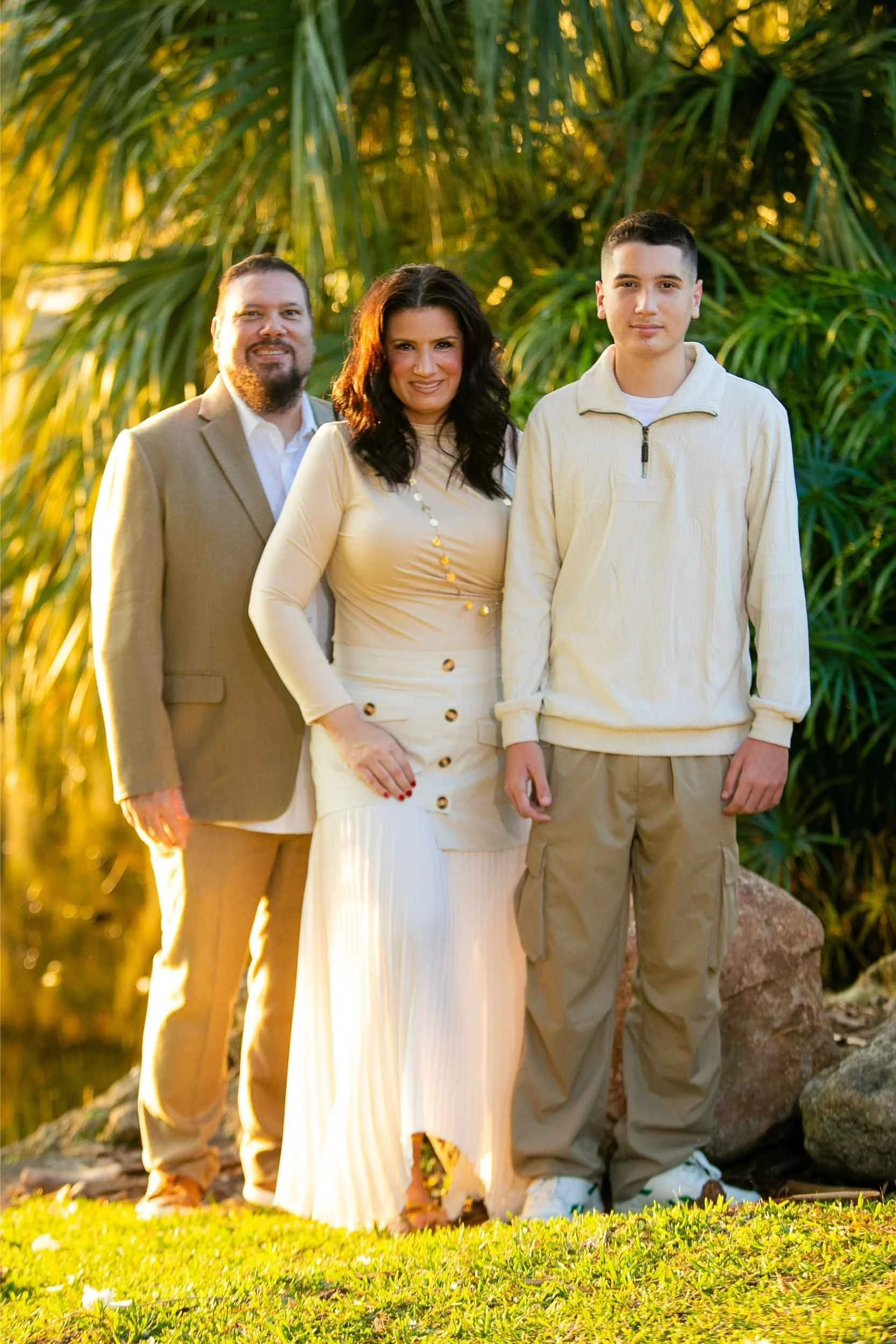 A family of three standing outdoors during golden hour, with tropical plants in the background, dressed in light-colored clothing.