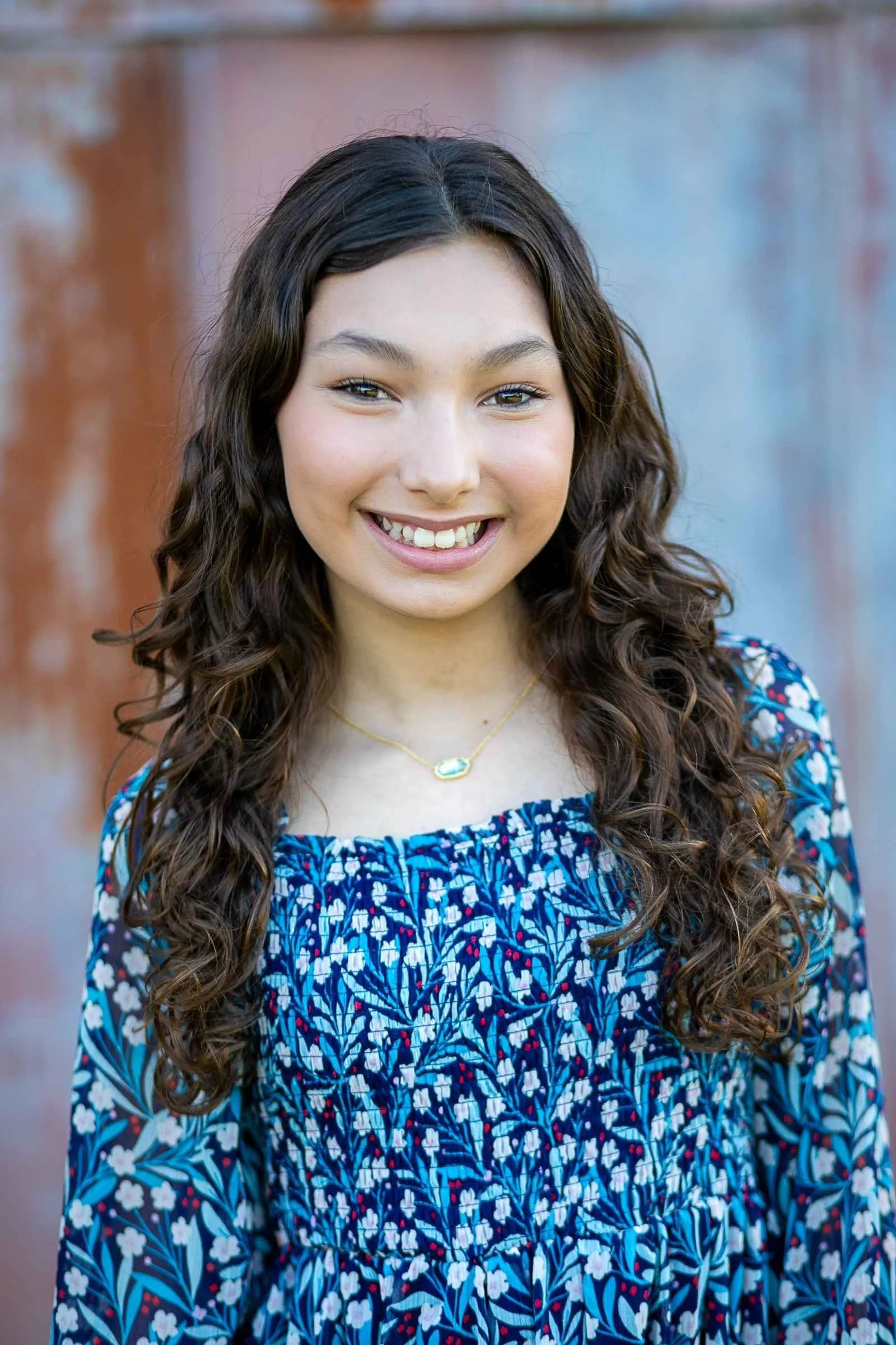 A young woman with long, curly brown hair smiling outdoors in front of a multicolored wall, wearing a blue floral dress and a gold necklace during a family portrait session with Cannonfire Photography.