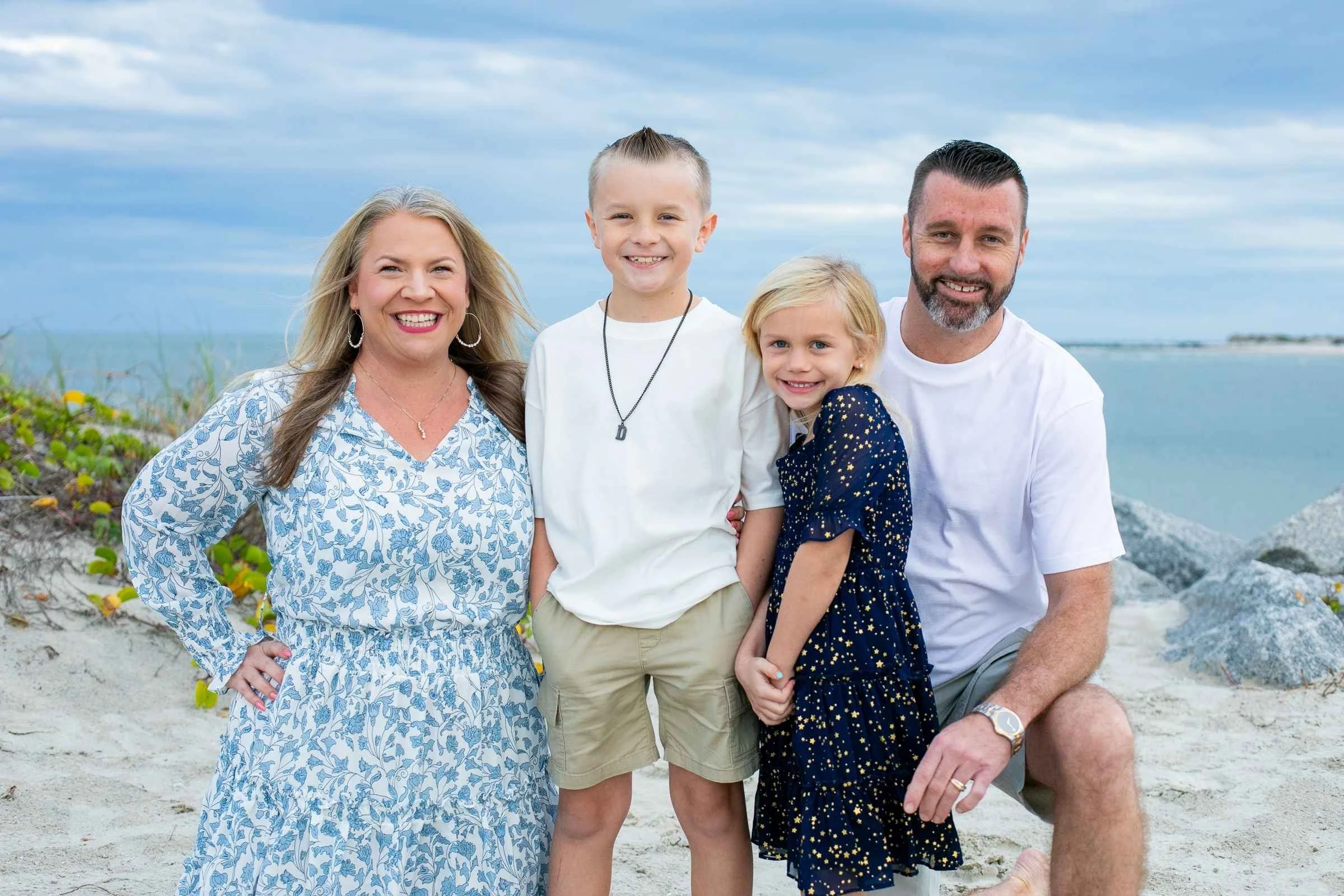 A family of four poses on the beach with sand, rocks, and sea in the background. The mother and father are kneeling on each side with their children in the middle. All are smiling and dressed in casual summer clothing.