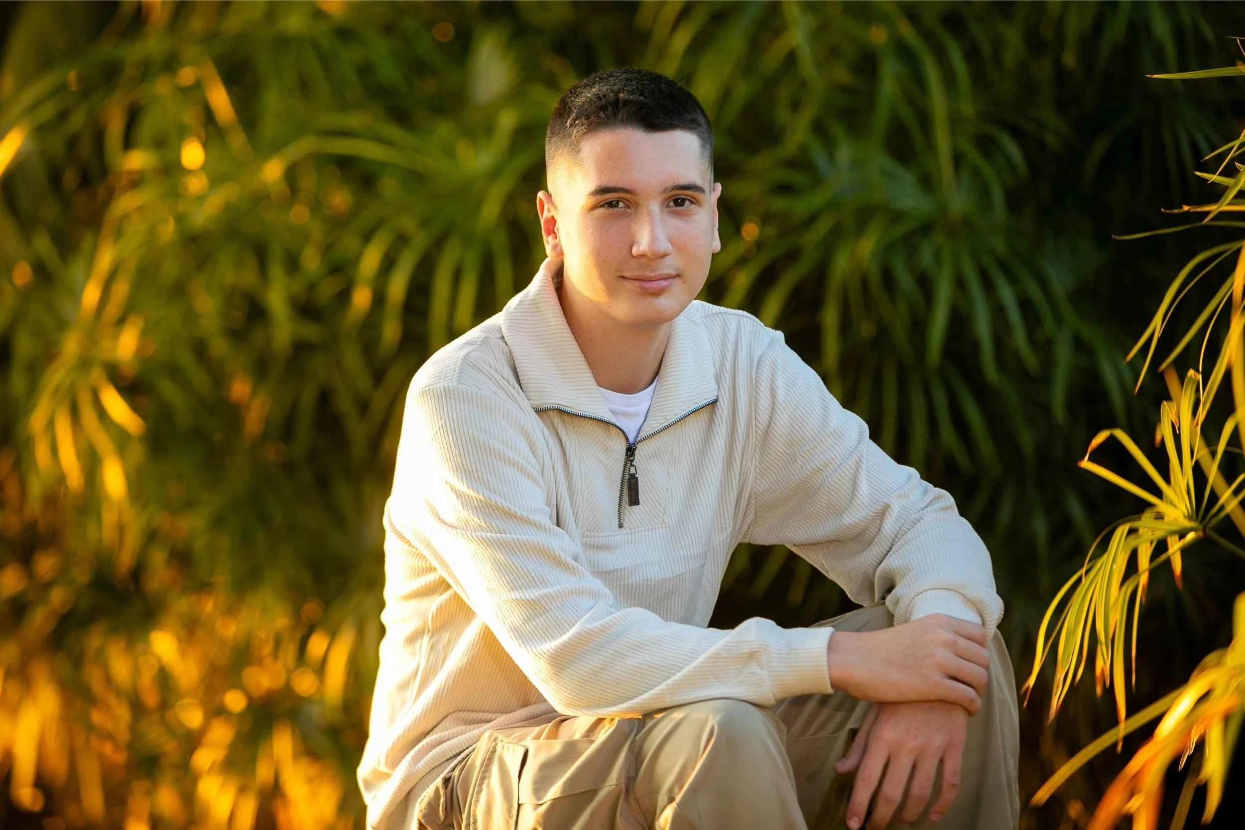 A young man sitting outdoors with greenery in the background, casually dressed in a cream sweater and beige pants, looking at the camera with a slight smile in downtown Orlando.