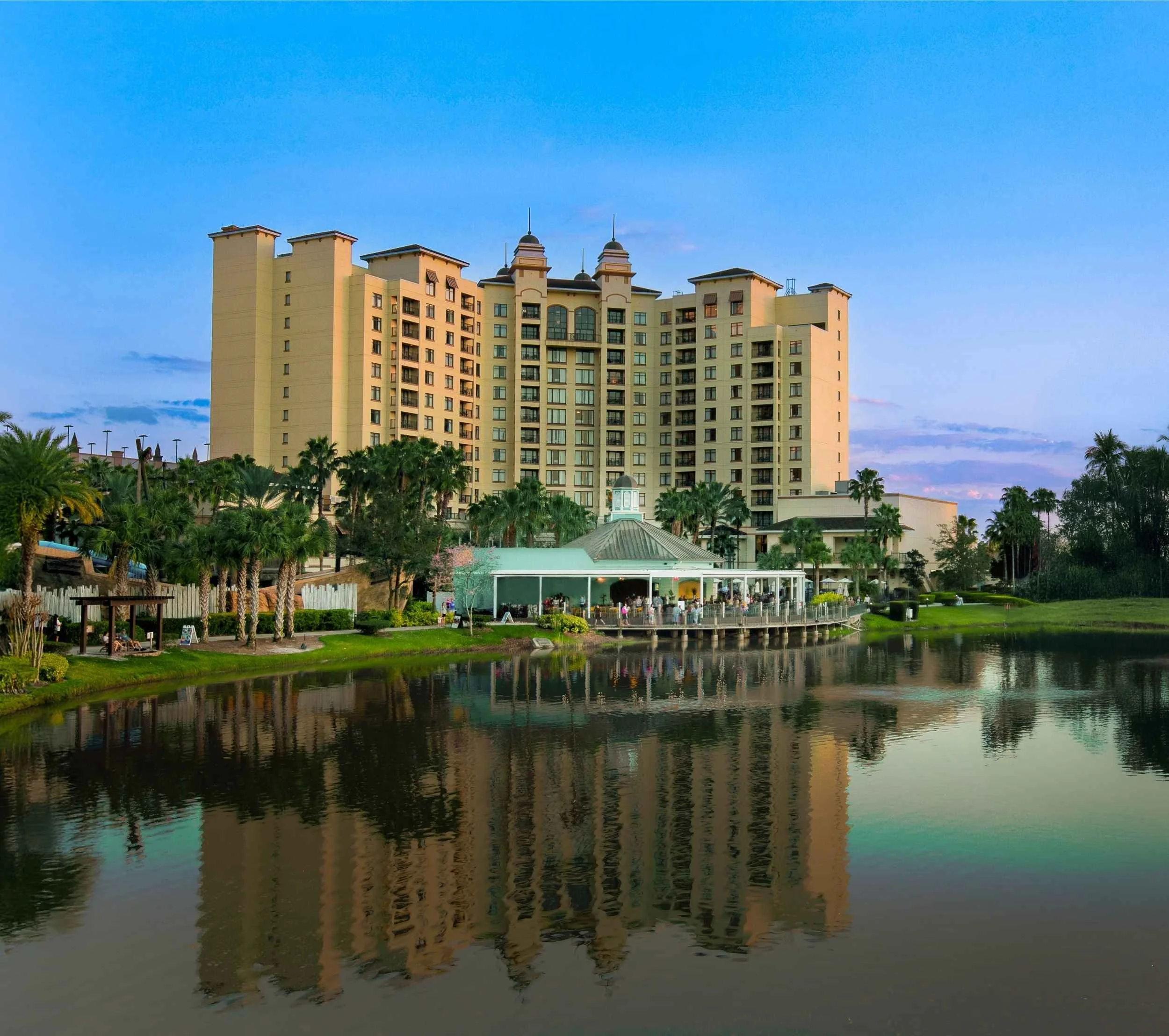 A large beige hotel building with multiple floors and towers, situated behind a pond with trees and a gazebo, during sunset.