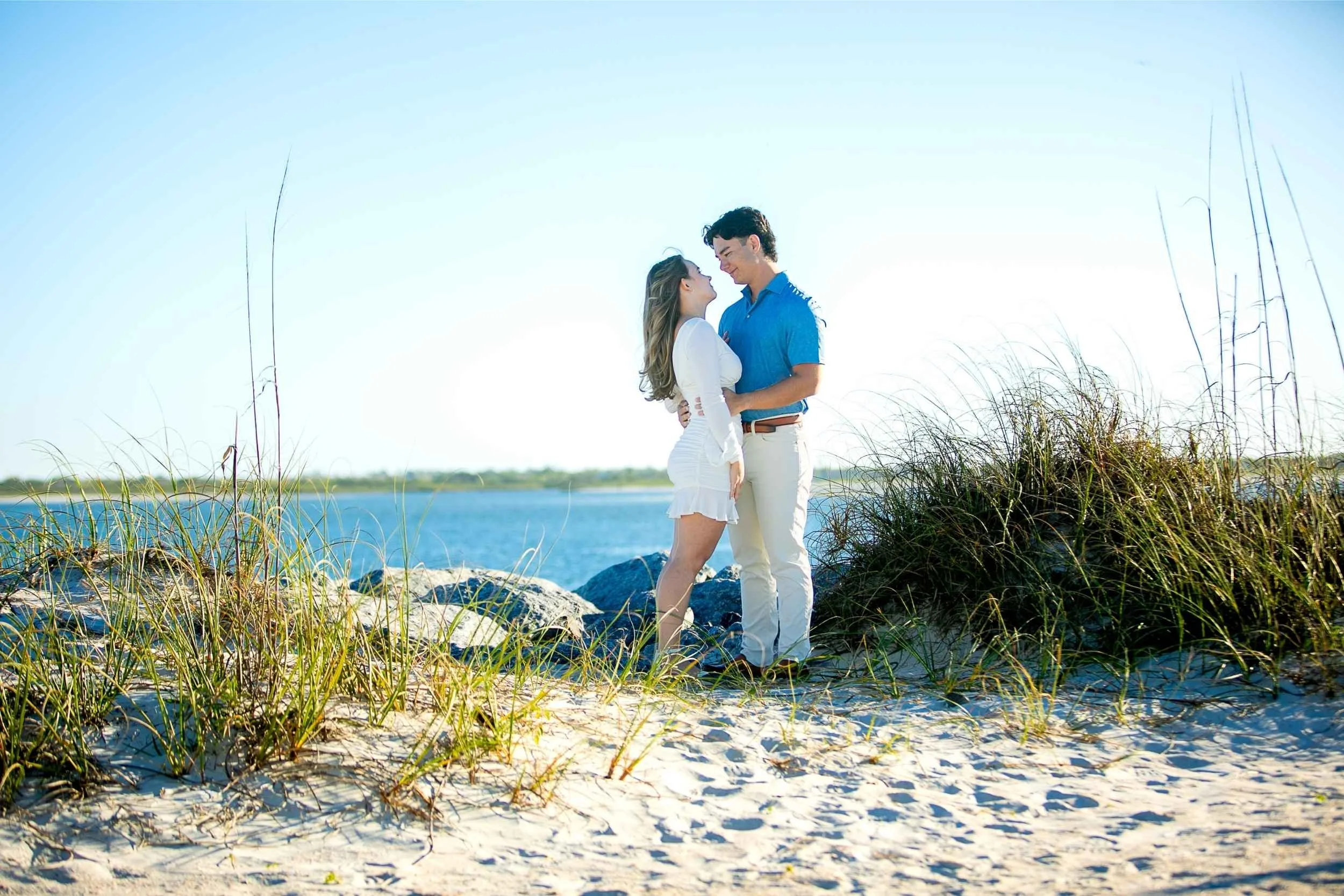 A young couple standing close together on a sandy beach by water, smiling and gazing into each other's eyes, with tall grass and rocks nearby under bright sunlight.