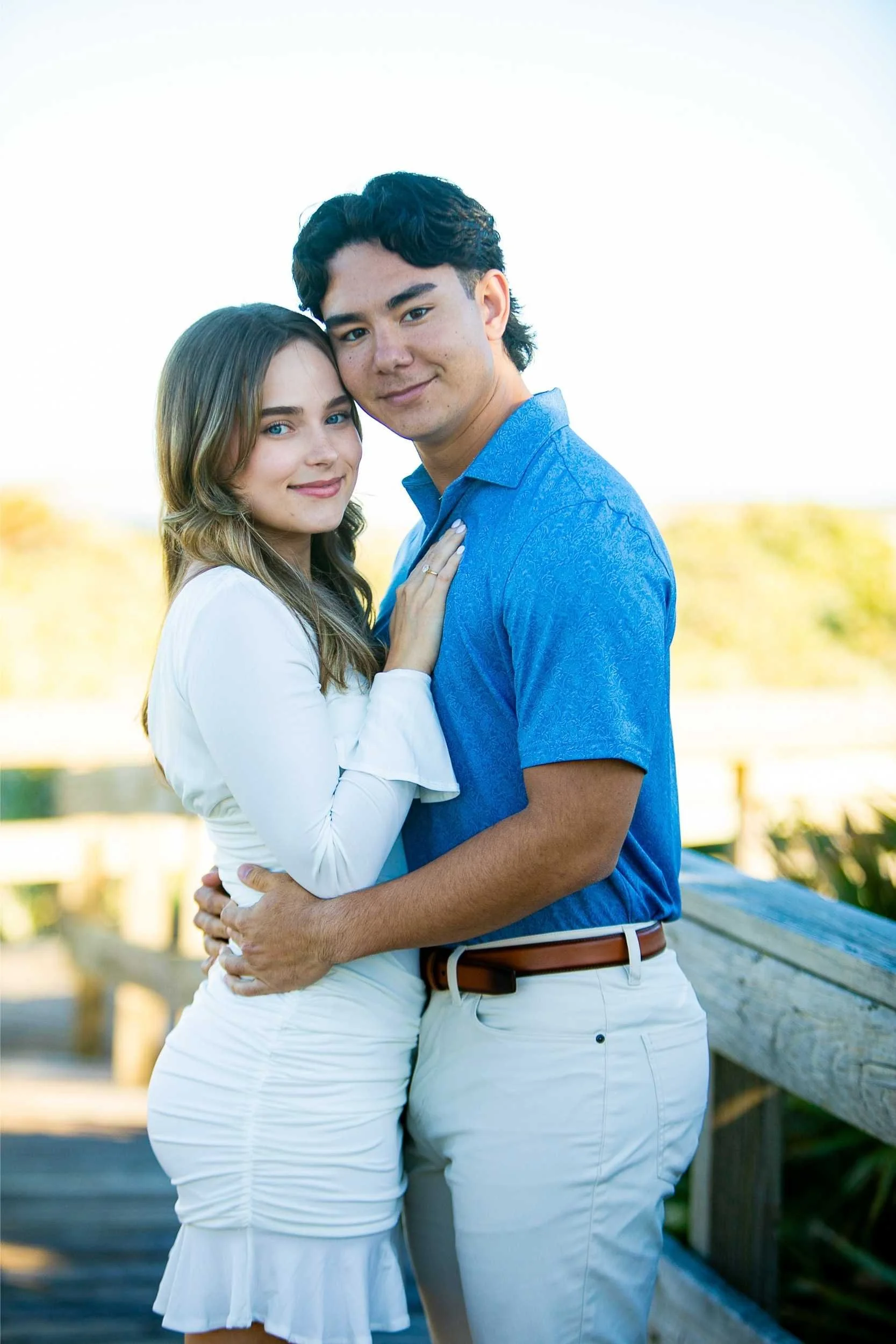 A young couple standing closely, smiling, outdoors with a blurred background of trees and a wooden fence, during daytime.