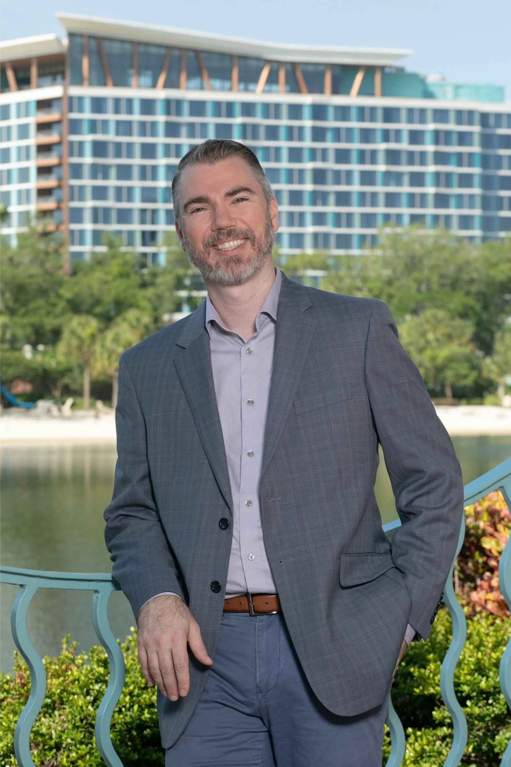 A man in a gray plaid blazer and light purple shirt standing outdoors on a bridge with a modern building and water in the background for a headshot with Cannonfire Photography.