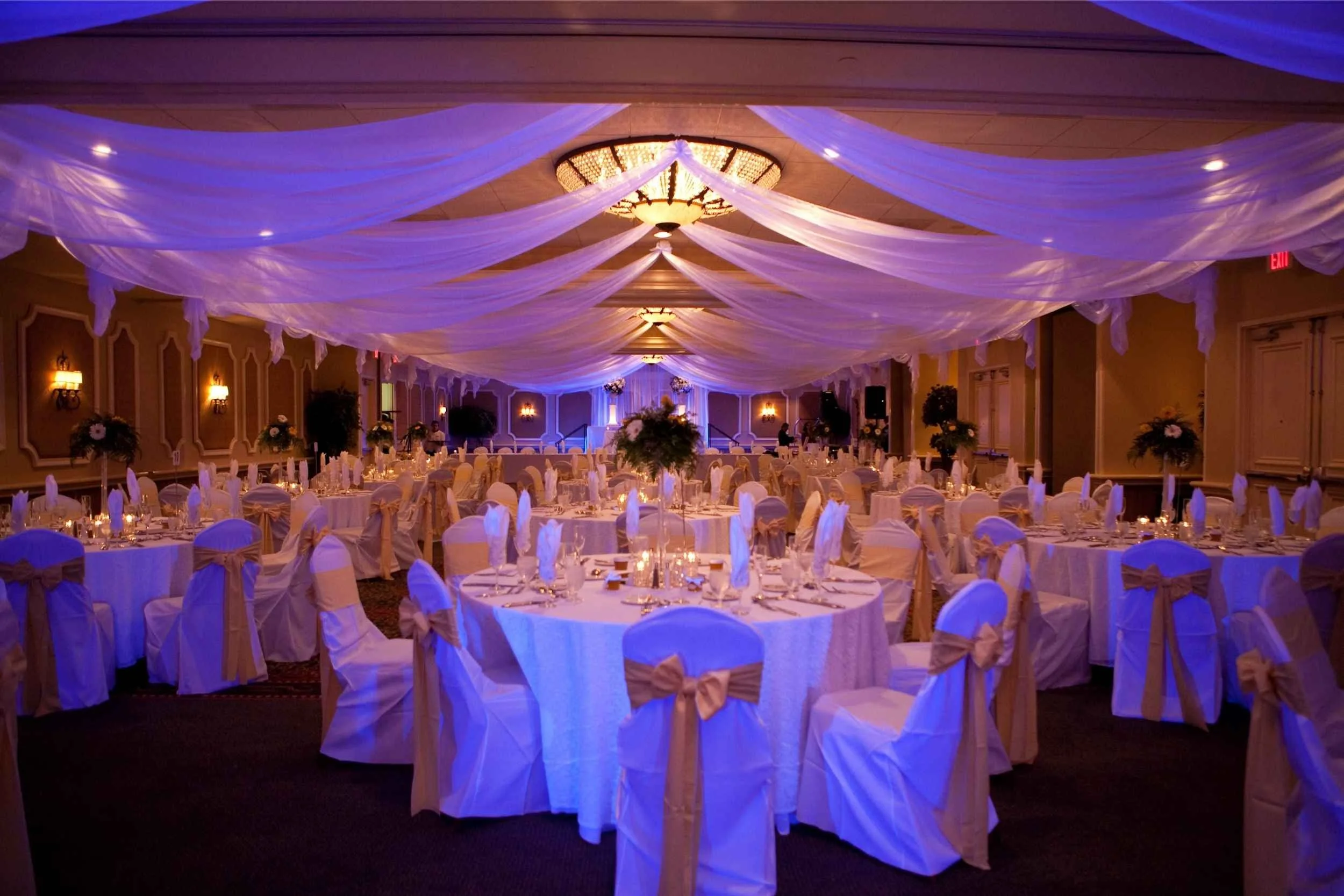 Elegant banquet hall decorated for an event, with draped white fabric on the ceiling, warm wall lighting, and round tables covered in white tablecloths and chairs with bows at a wedding photographed by Cannonfire Photography.