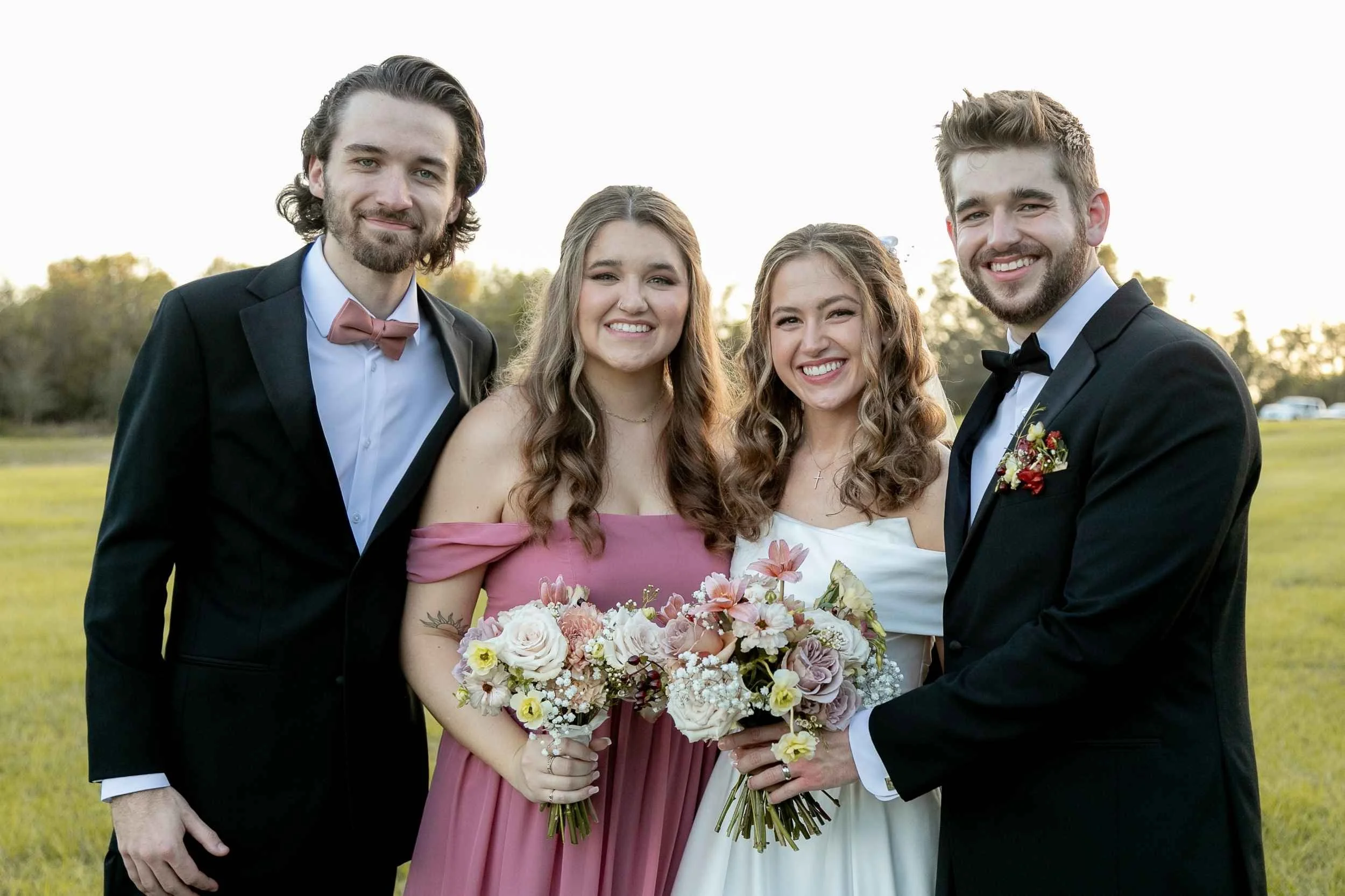 Four people dressed in formal attire smiling outdoors during a wedding, holding bouquets of flowers for a wedding photographed by Cannonfire Photography.