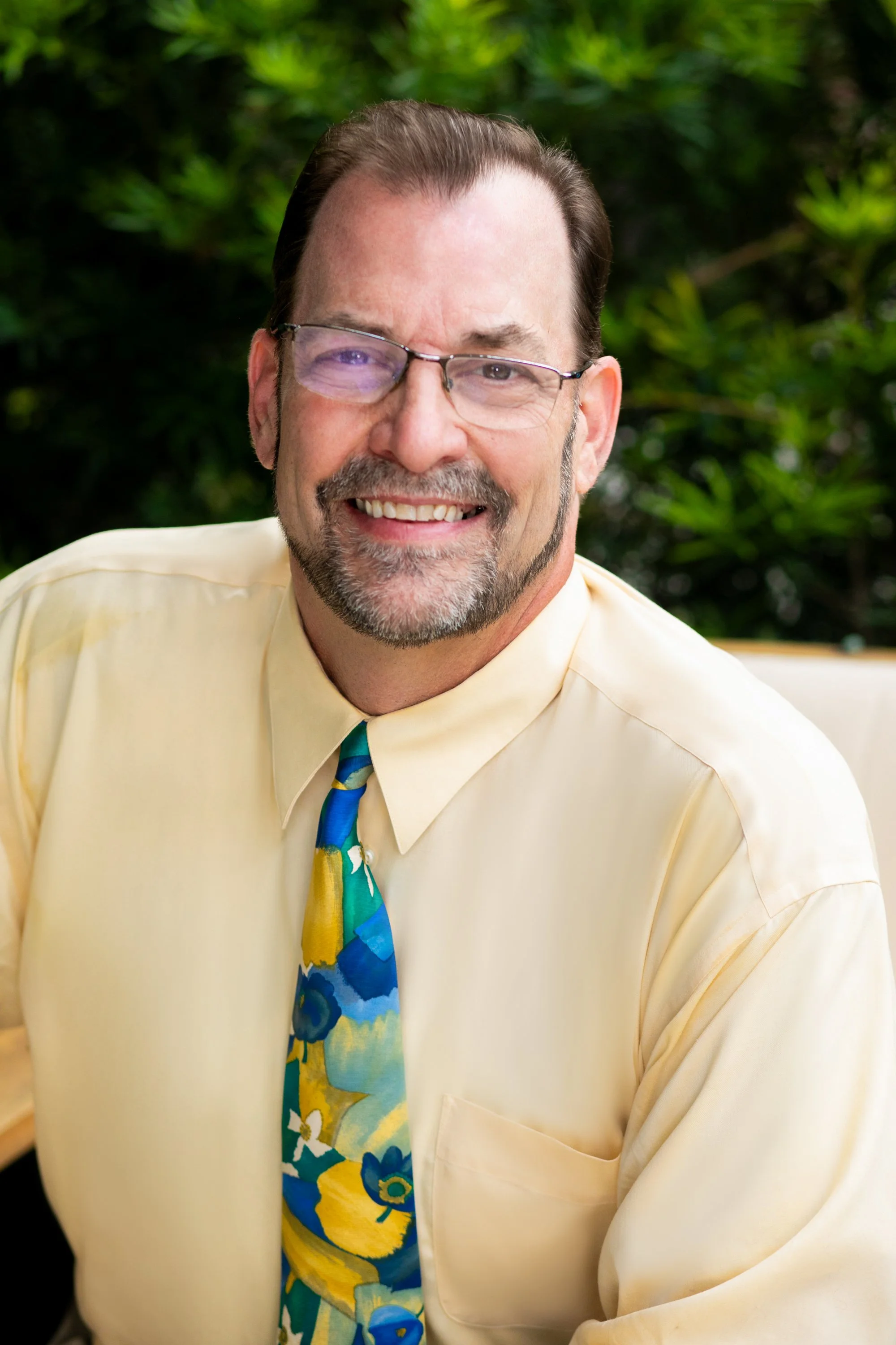 Portrait of a smiling man with glasses, facial hair, wearing a light yellow shirt and a colorful blue and yellow tie, sitting against a green outdoor background for a headshot with Cannonfire Photography in Orlando.
