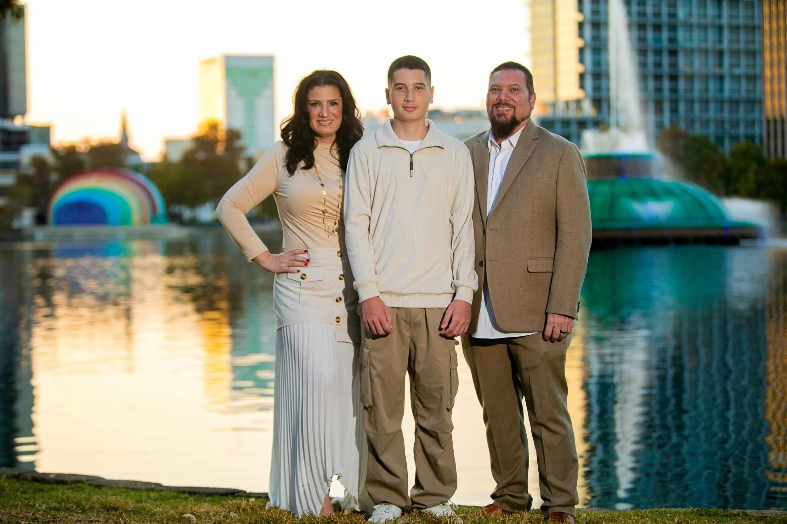 A family of three standing outdoors in front of a lake and cityscape during sunset, with buildings and a fountain in the background. The mother is wearing a beige dress, the son is wearing a cream-colored sweater and khakis, and the father is wearing