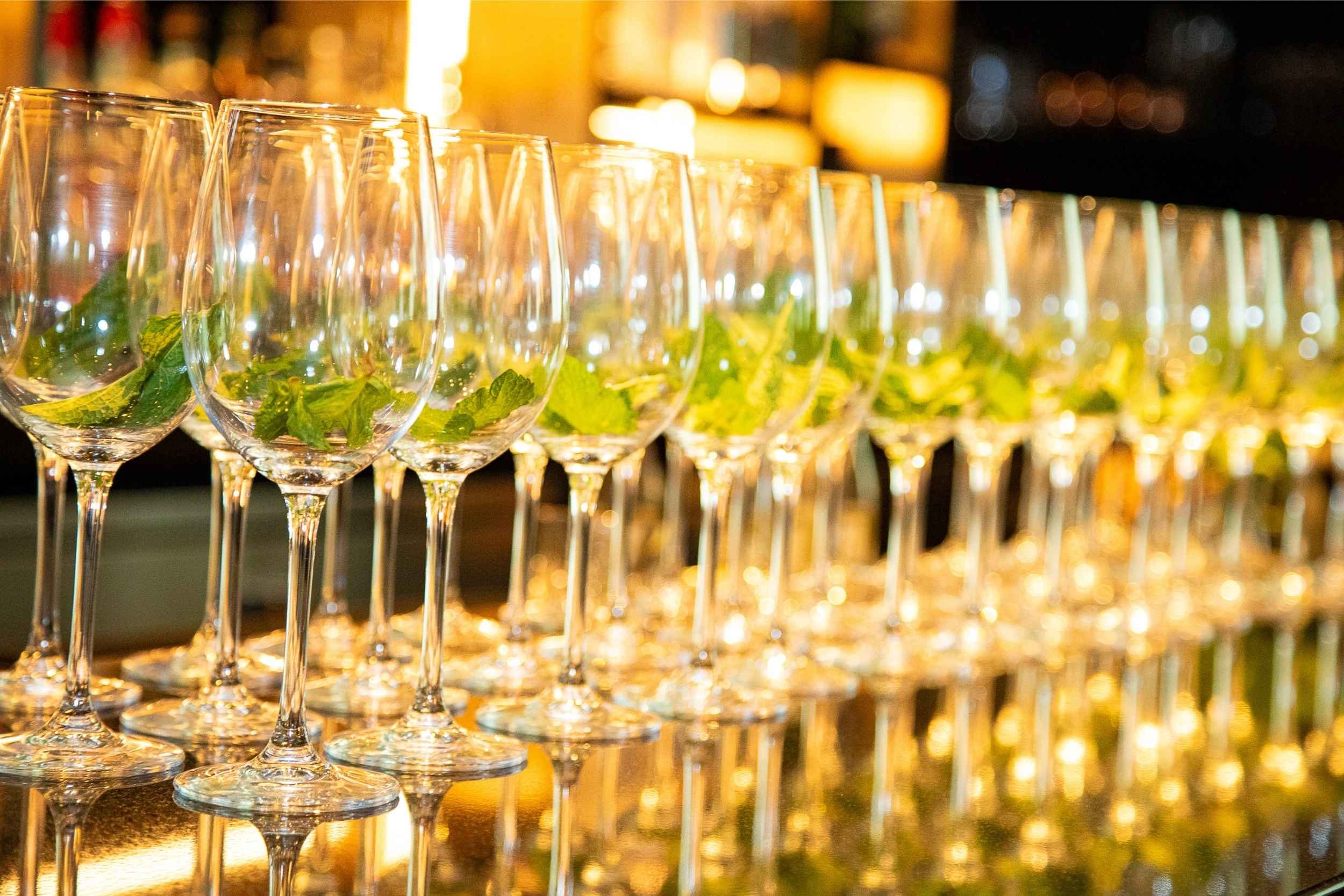 Row of empty champagne glasses with green mint leaves inside, reflected on a shiny surface, with warm, blurry lights in the background.