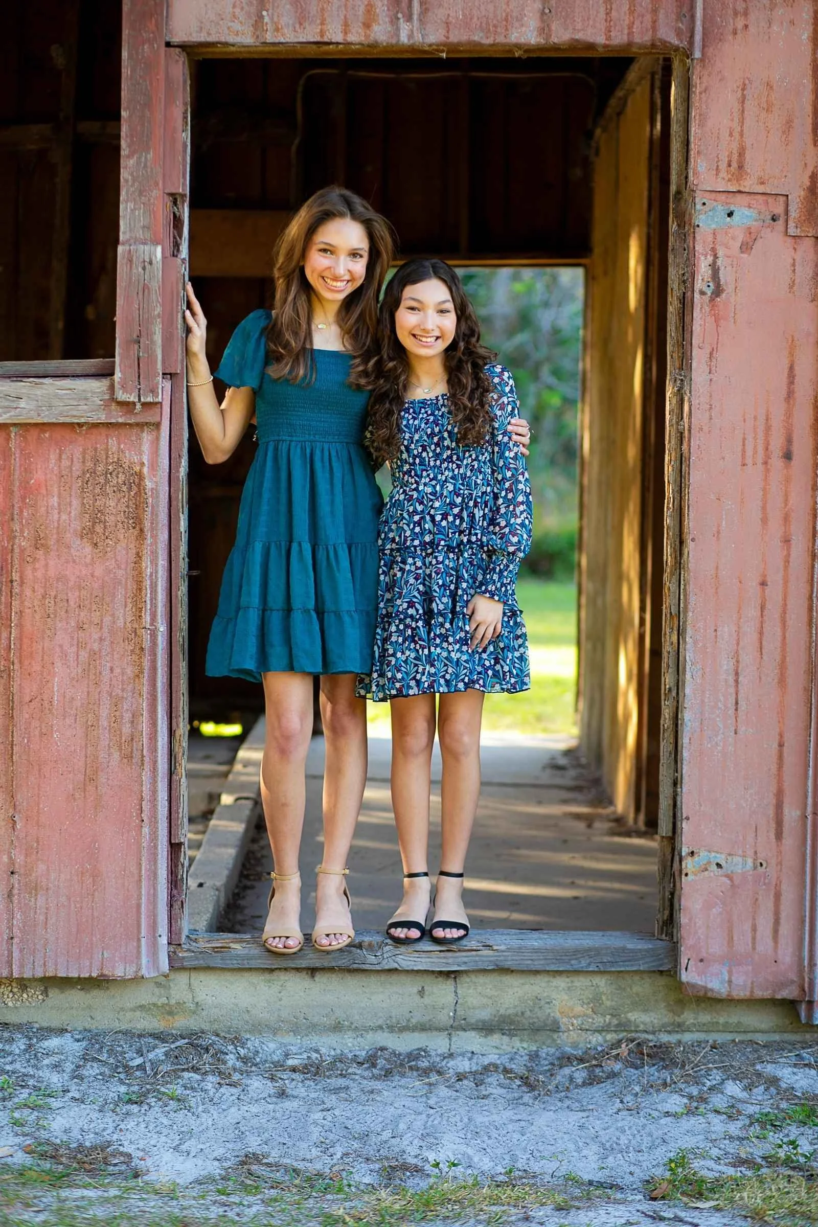 Two young women in dresses standing together on a wooden porch of an old rustic barn, smiling at the camera with greenery in the background for a family portrait session with Cannonfire Photography.