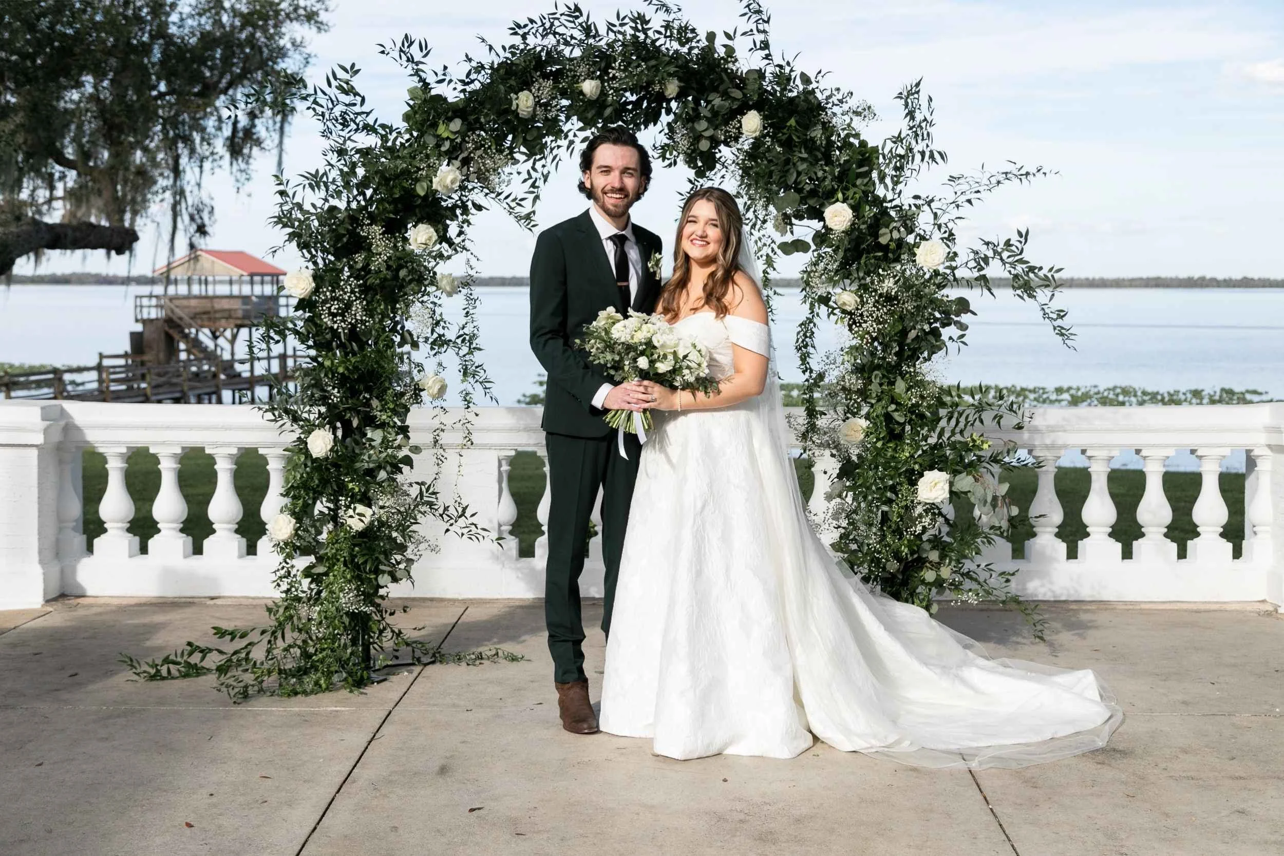 A bride and groom standing together under a green and white floral arch on an outdoor deck, with water and a dock visible in the background. The bride is wearing a white off-the-shoulder wedding dress holding a bouquet, and the groom is dressed in a 