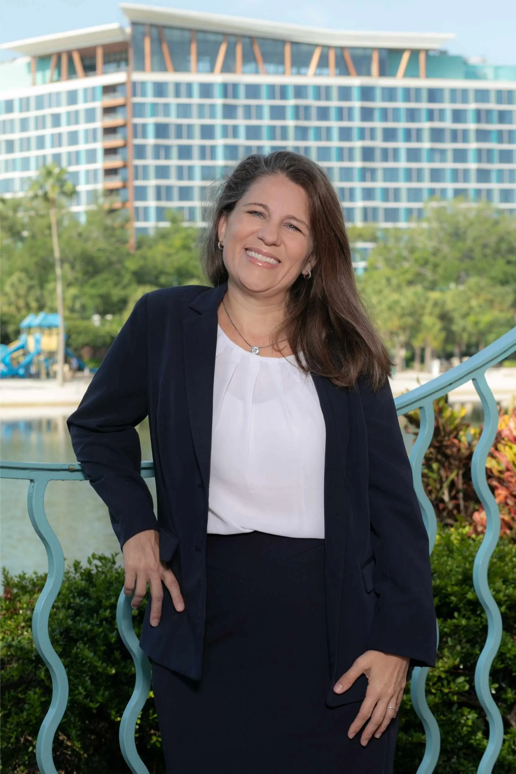 A woman in business attire smiling outdoors, standing next to a blue railing with a modern building, trees, and a body of water in the background for a headshot with Cannonfire Photography.