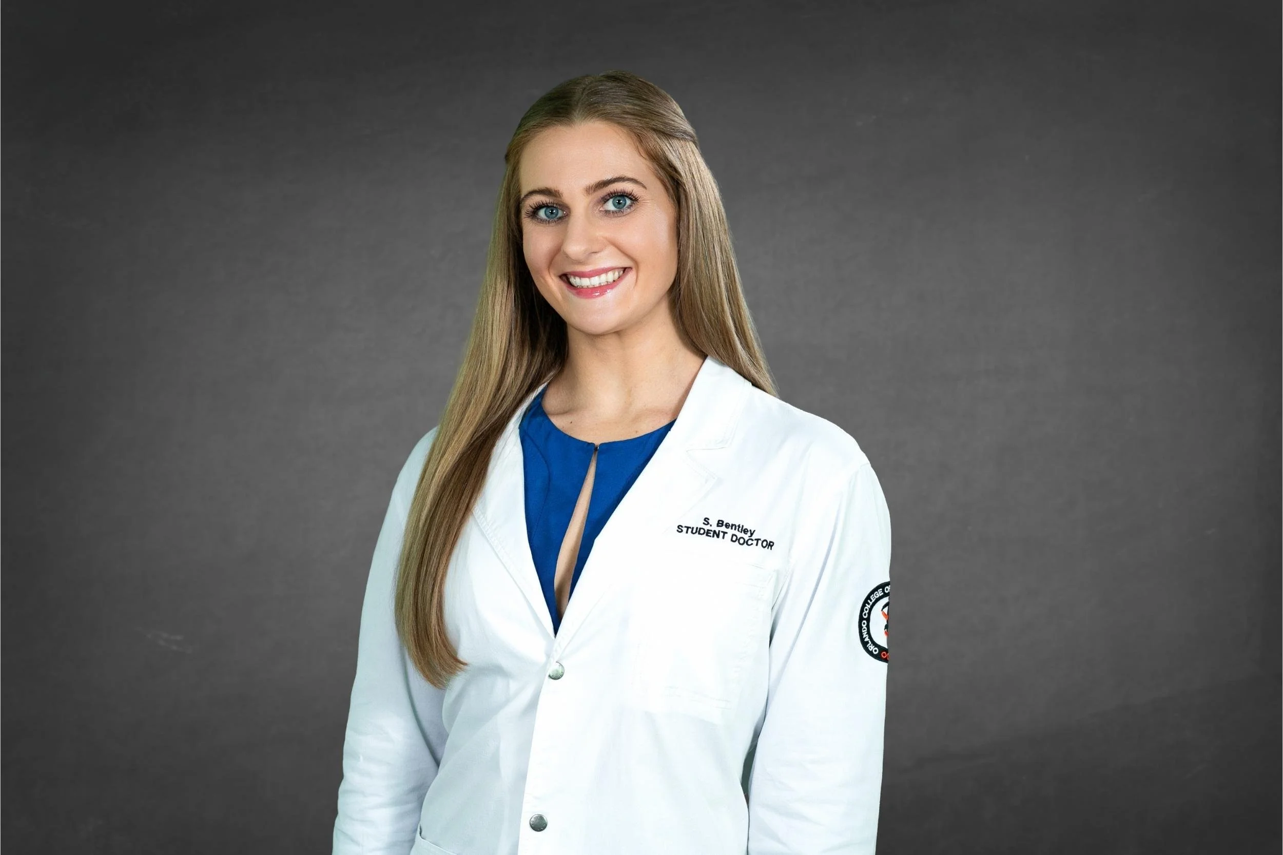 Portrait of a young female medical professional in a white coat with a name tag, standing against a gray background for a headshot with Cannonfire Photography in Orlando.