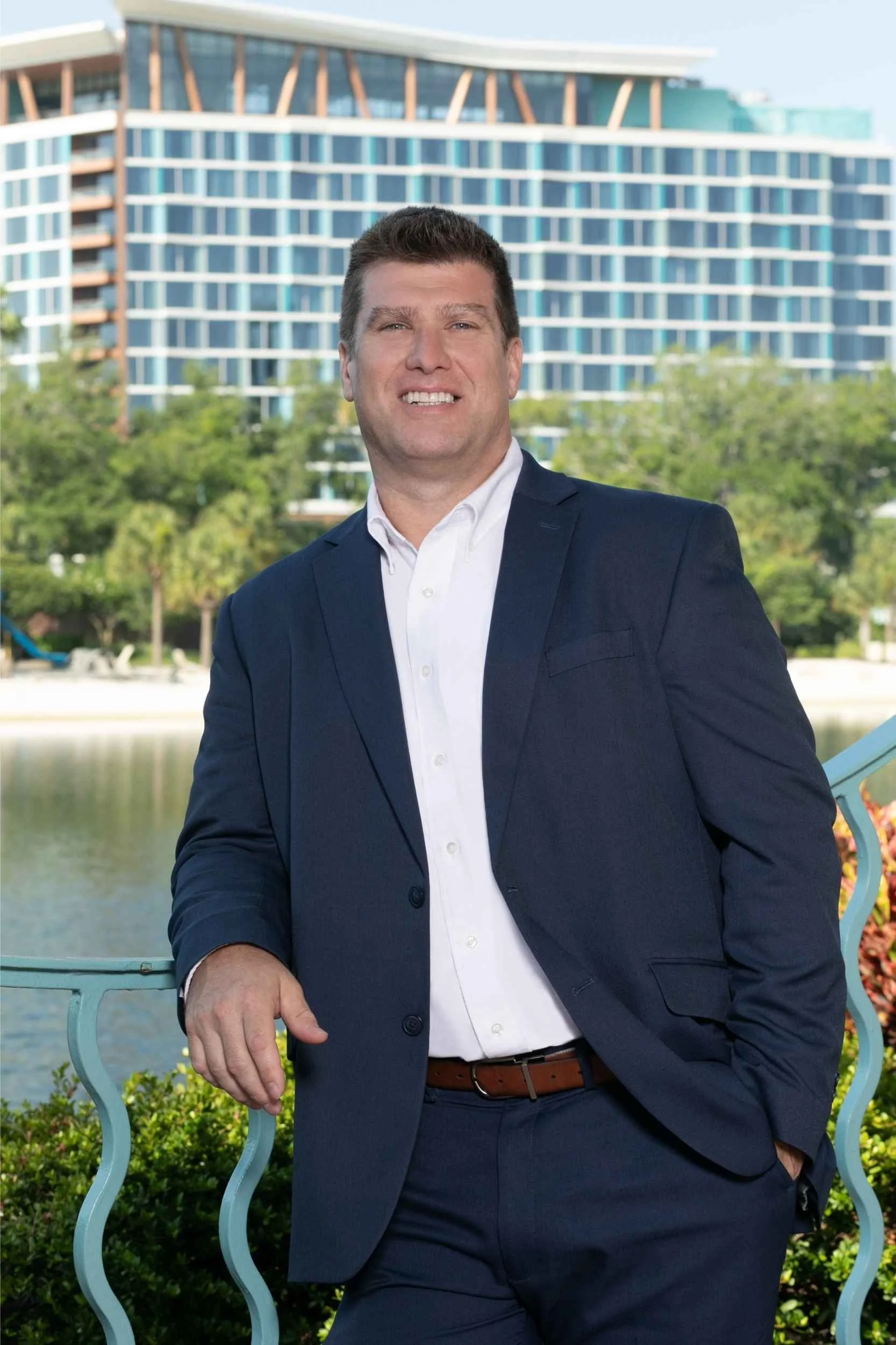 A man in a navy suit and white shirt standing outdoors near a body of water with a modern high-rise building in the background for a headshot with Cannonfire Photography.
