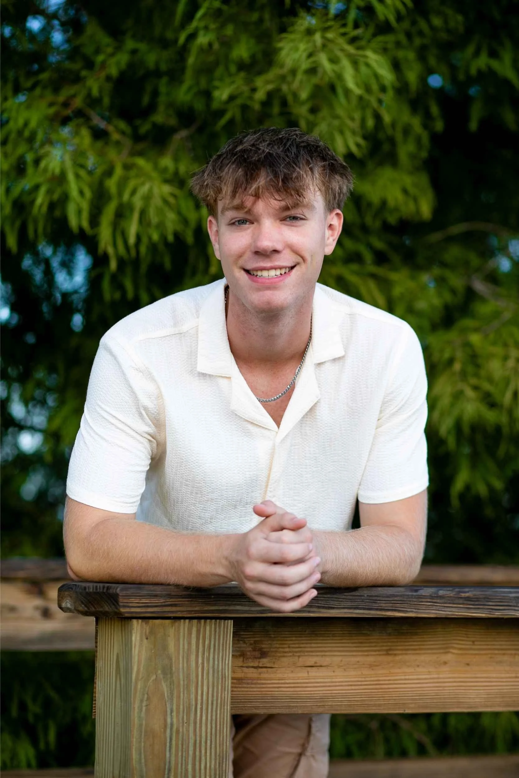 Young man with brown hair wearing a white shirt, smiling, leaning on a wooden railing outdoors for a senior portrait session with Cannonfire Photography.