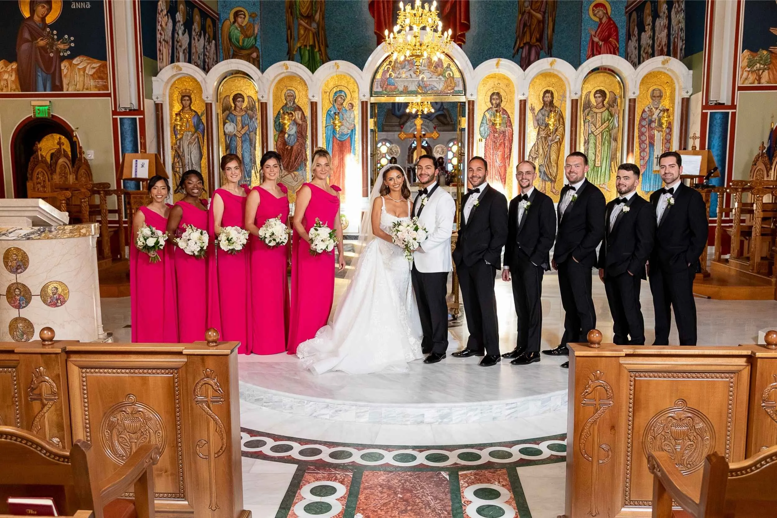 A wedding party standing inside a church, with the bride and groom at the center. The church features religious icons and artwork on the walls, with warm lighting. The bridal party includes five bridesmaids dressed in pink, holding white bouquets, an