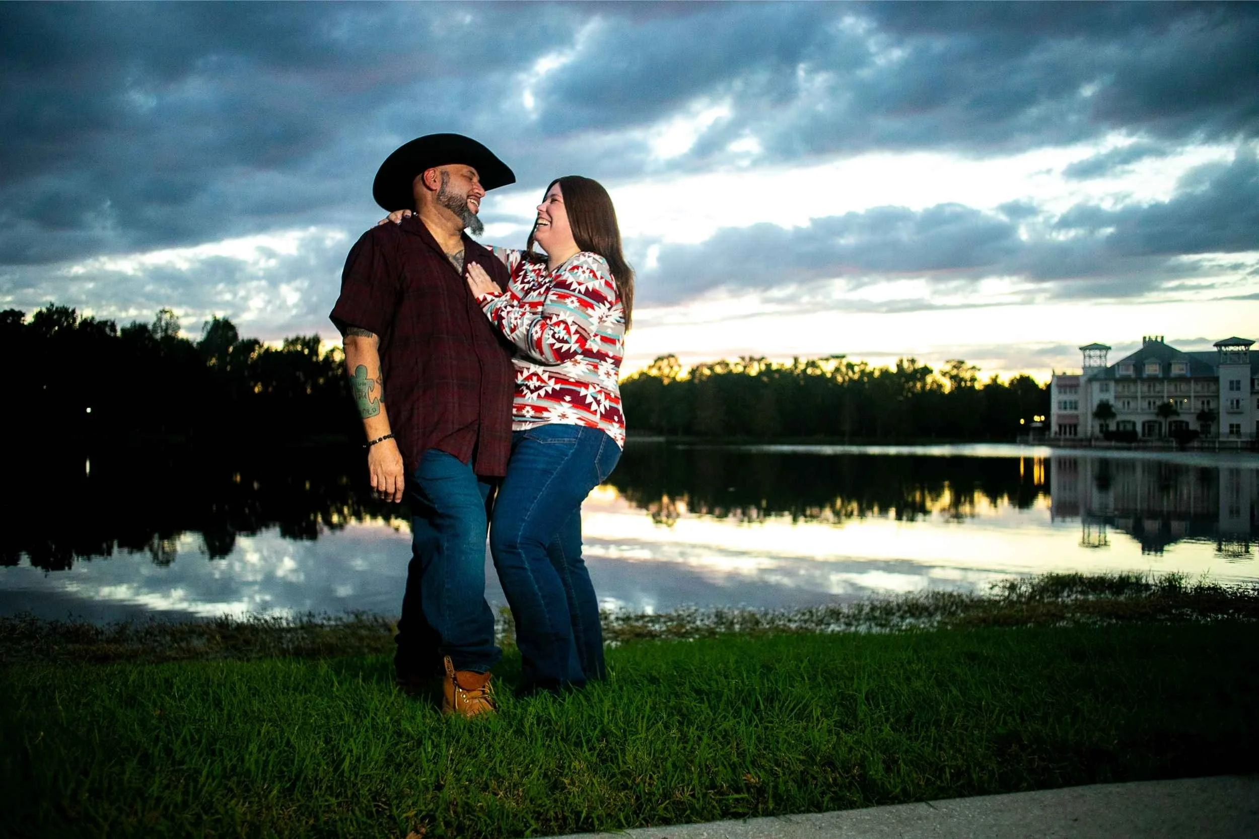 A smiling man and woman standing near a lake at sunset, embracing and looking at each other. The man wears a cowboy hat and plaid shirt, and the woman wears a colorful patterned sweater. A large house is visible in the background.