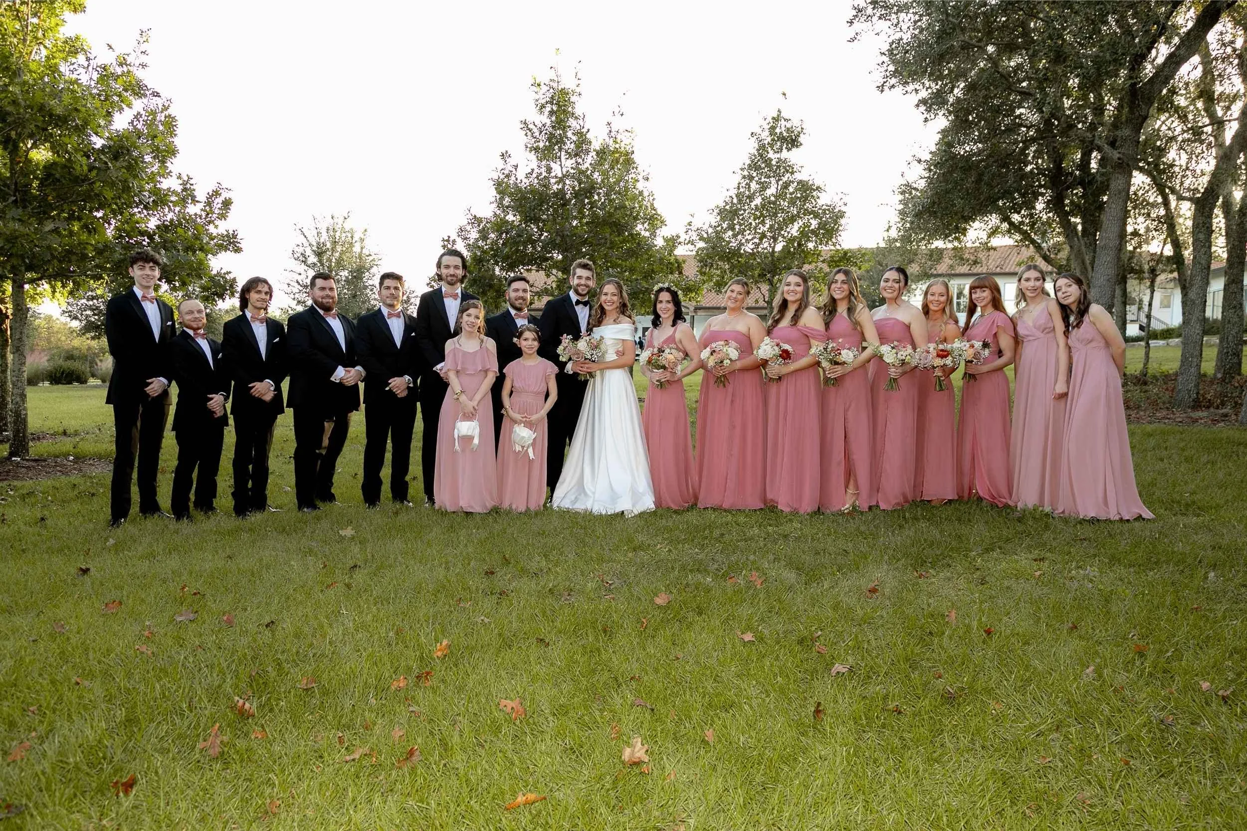 A large wedding party outdoors on grass, with men in black tuxedos and women in pink dresses, standing under trees for a wedding photographed by Cannonfire Photography in Lake Wales.