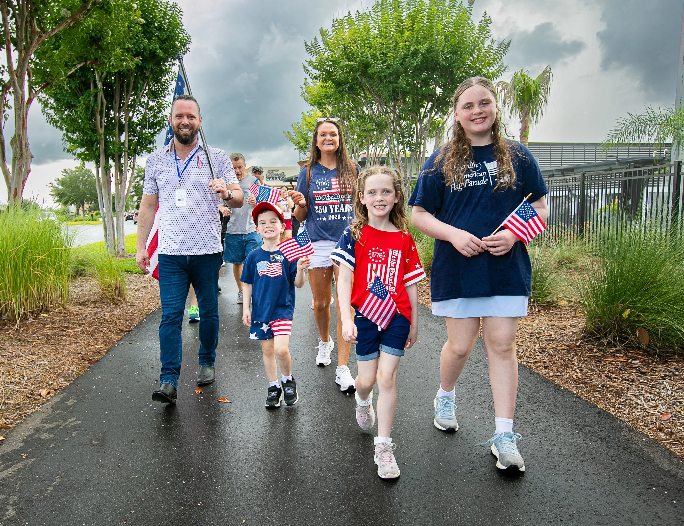 A group of people, including children and adults, are walking outdoors on a sidewalk decorated for a patriotic celebration, carrying small American flags. The sky is cloudy, and there are trees and a fence in the background.