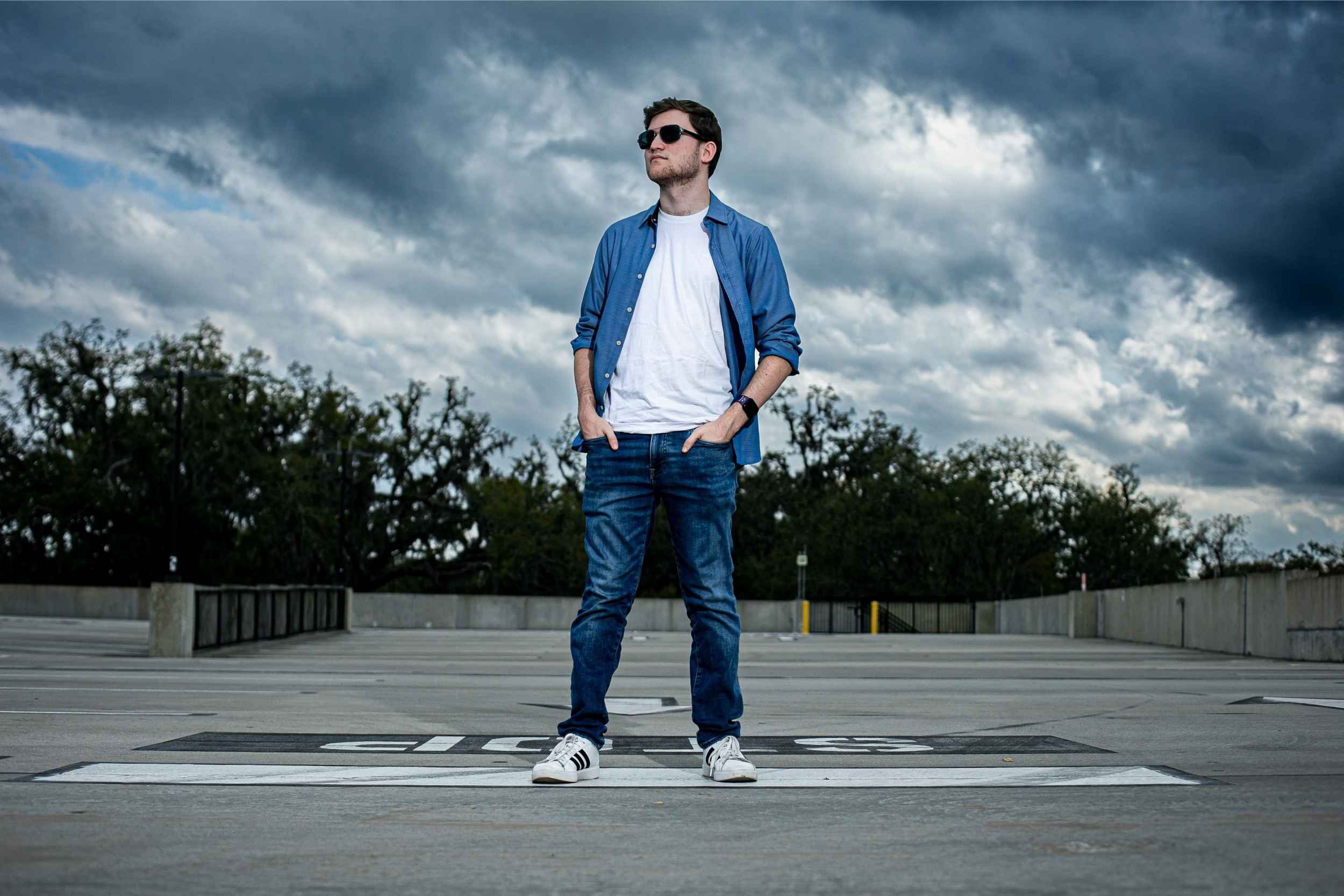 Young man standing on a parking lot with a cloudy sky in the background, wearing sunglasses, a white t-shirt, blue jeans, and a blue open shirt for a senior portrait session with Cannonfire Photography.