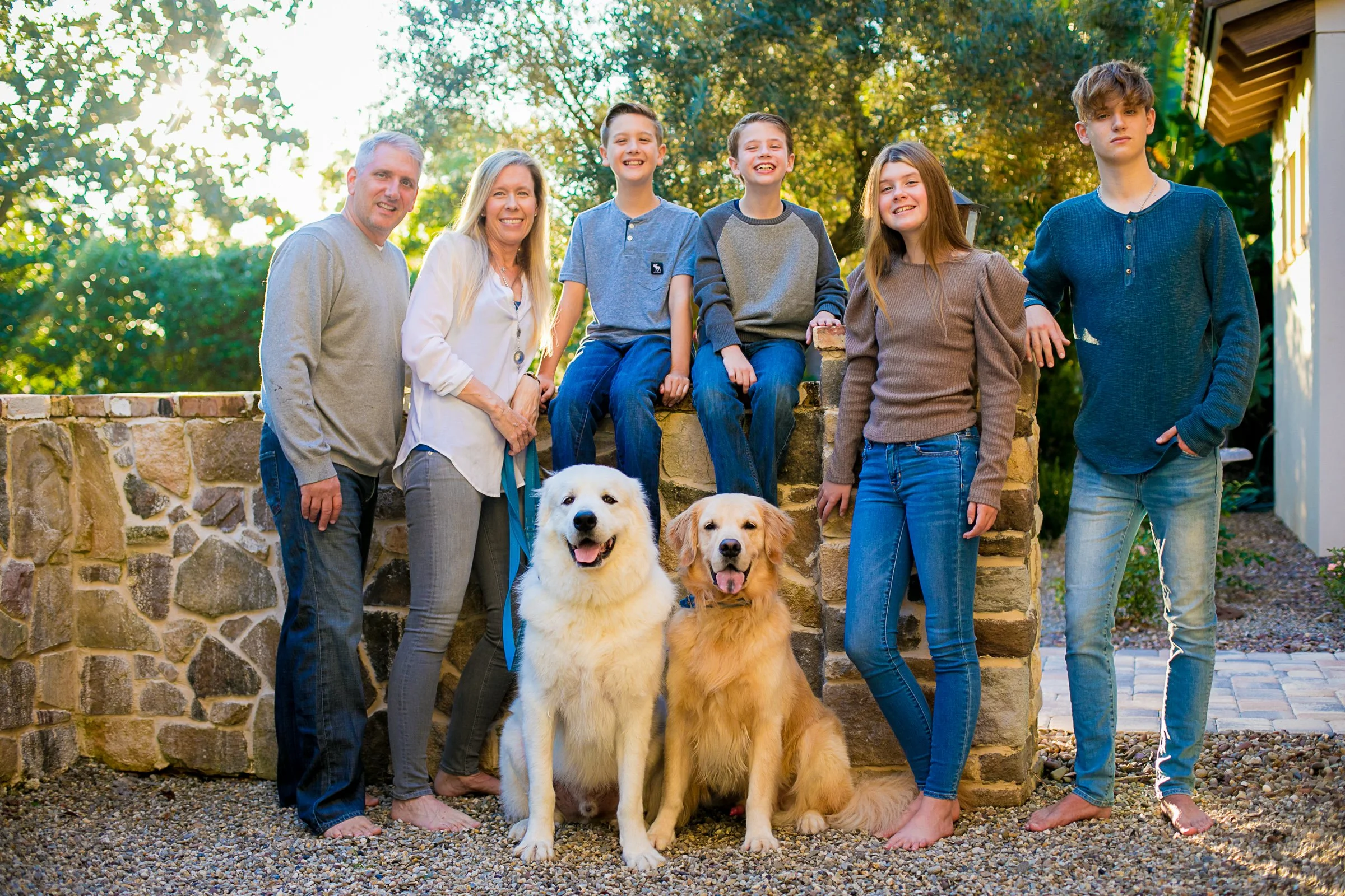 A family of seven, including two adults, four children, and two dogs, standing outdoors on a gravel area in front of a stone wall with trees and sunlight in the background for a family portrait with Cannonfire Photography in Orlando.