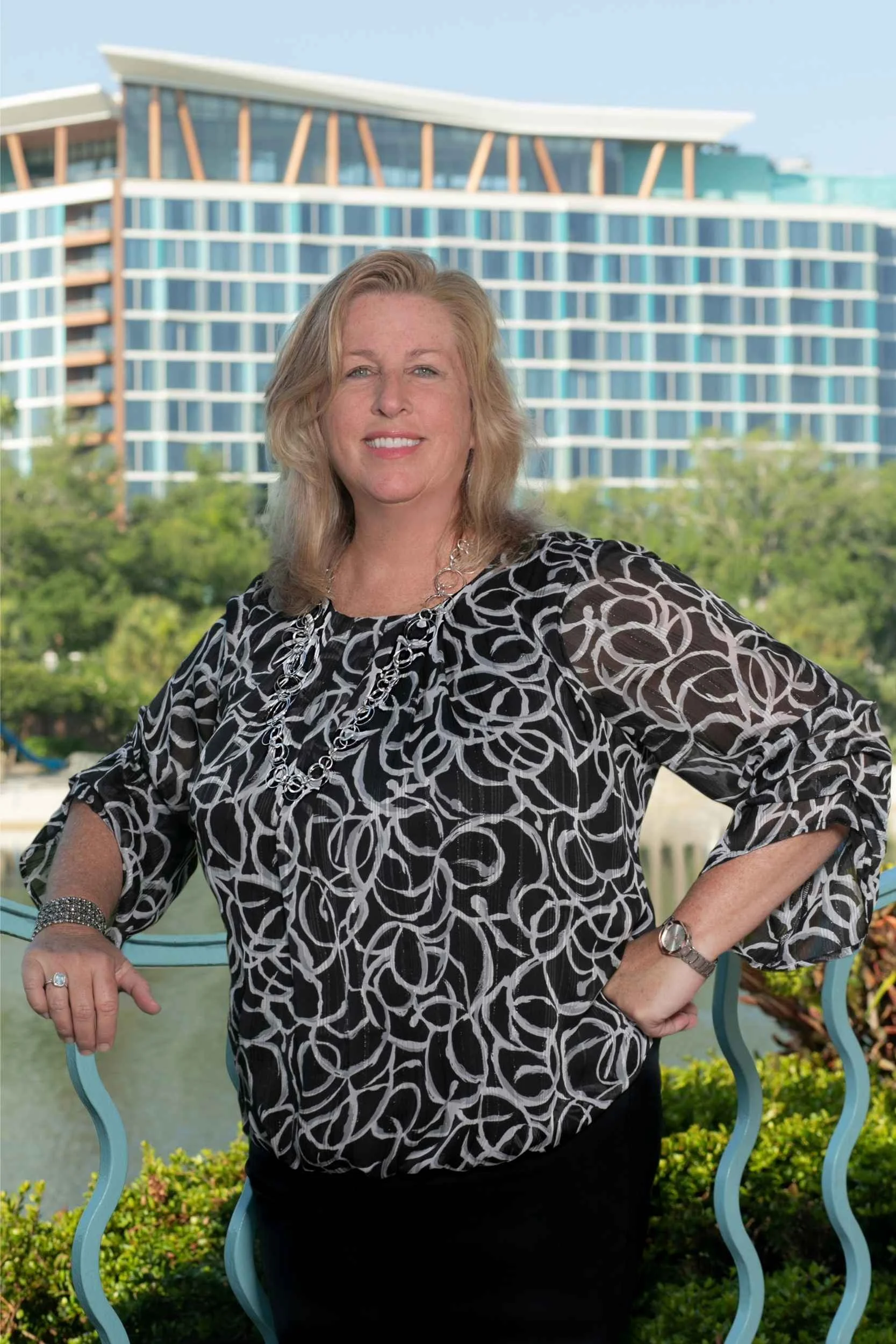 A woman standing outdoors on a balcony with a modern building and trees in the background. She has blonde hair, is smiling, and is wearing a black and white patterned top with jewelry for a headshot with Cannonfire Photography.