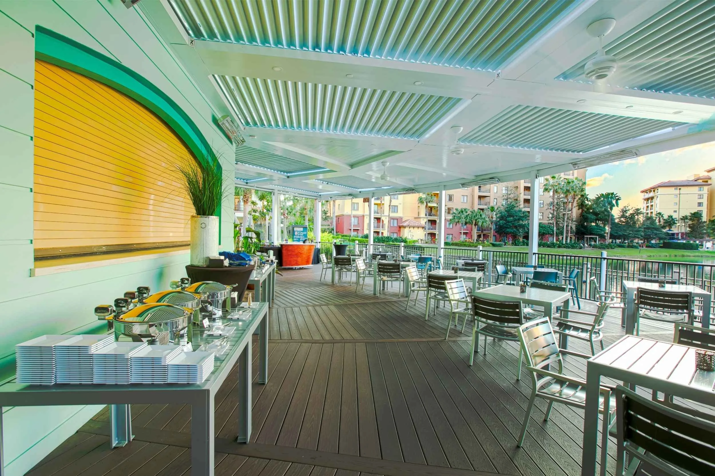Outdoor dining area with tables, chairs, and a buffet table, overlooking a sunny courtyard with palm trees and apartment buildings in the background.