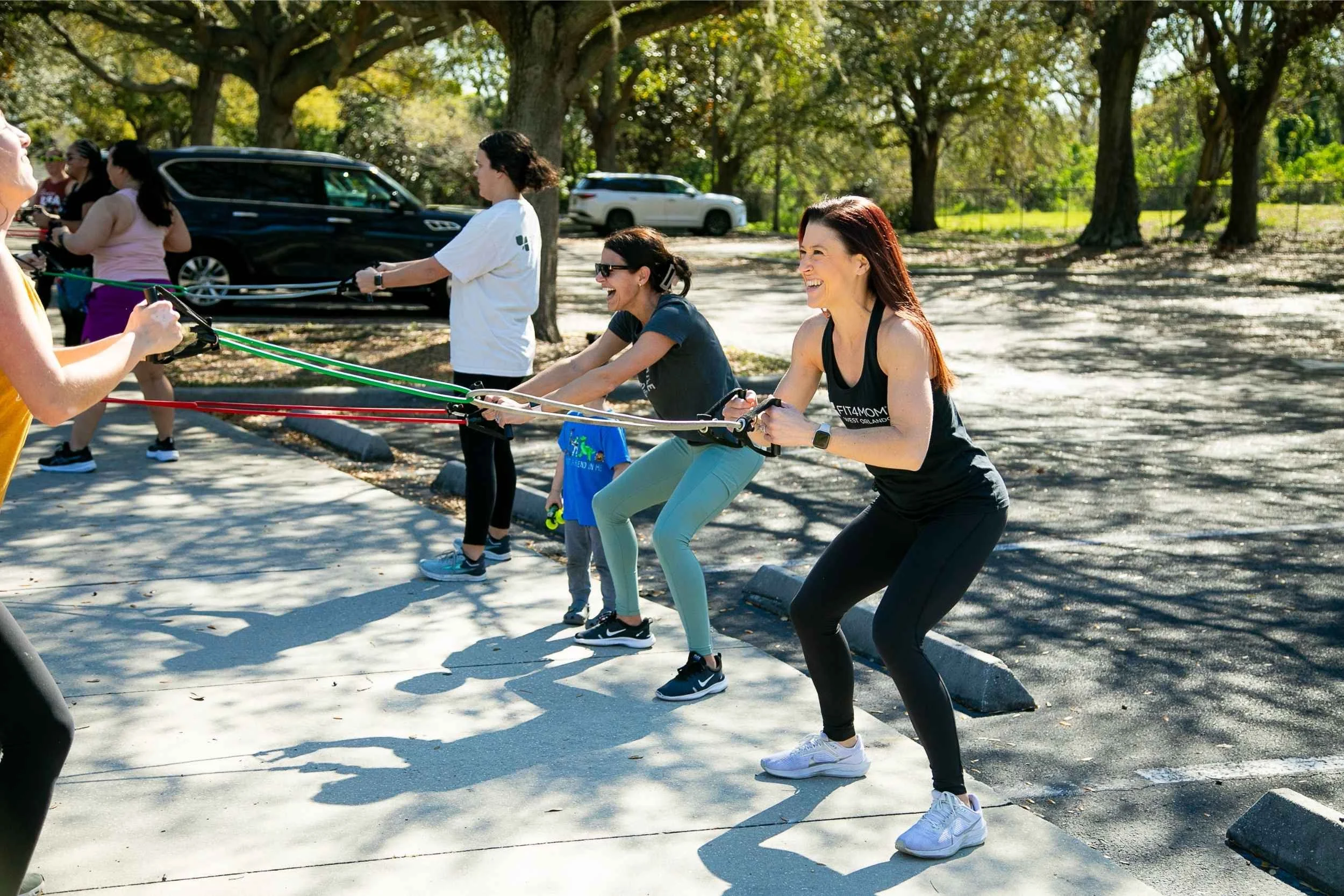 Group of women participating in an outdoor fitness class pulling resistance bands, smiling, wearing workout clothes, in a park with trees and parked cars for an advertising photography session by Cannonfire Photography in Winter Garden.