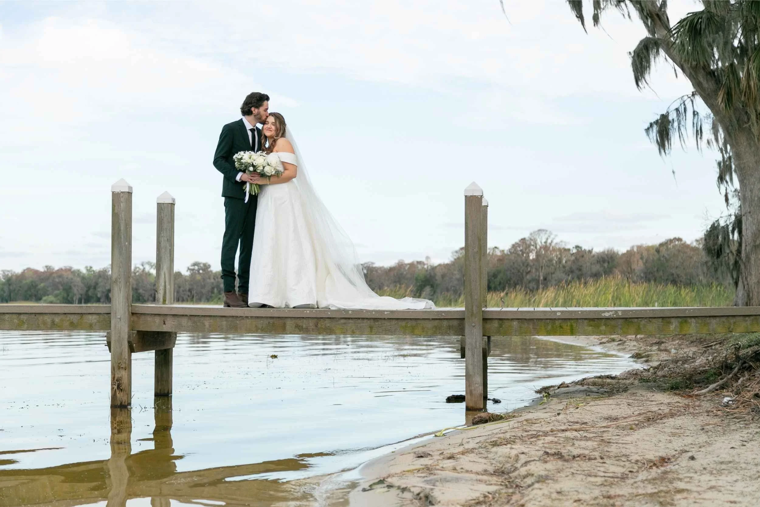 A bride and groom standing on a wooden dock near water, embracing, with a bride holding a flower bouquet, outdoors against a natural landscape with trees and sky for a wedding photographed by Cannonfire Photography.