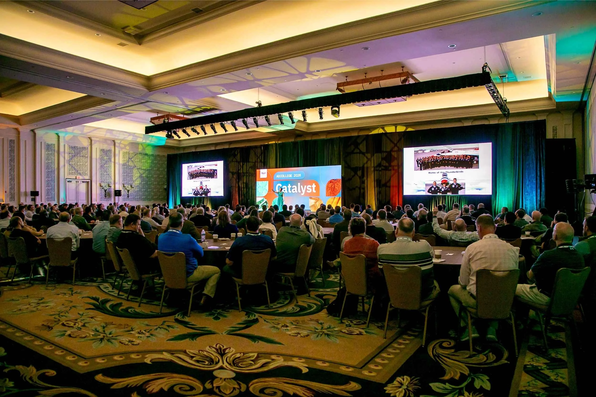 Conference hall filled with attendees, large stage with screens showing a presentation titled 'Catalyst,' and people seated at round tables for a conference.