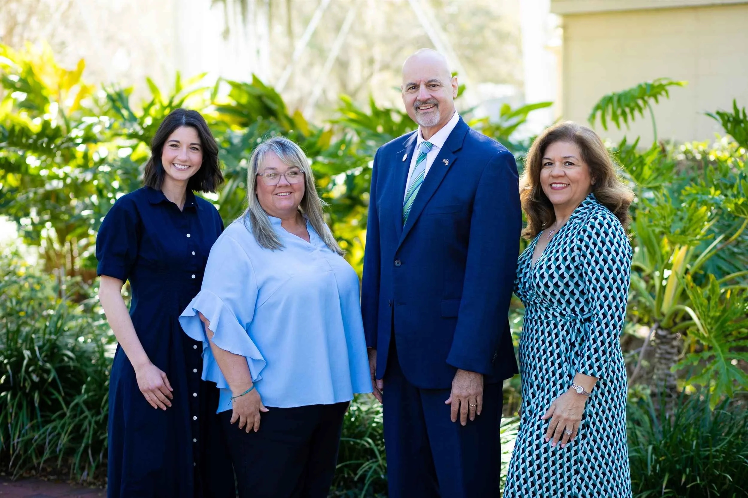 Four people standing outdoors in front of green foliage. The group includes three women and one man, all smiling and dressed in professional attire. The man is wearing a blue suit and tie, and the women are in dresses or blouses.
