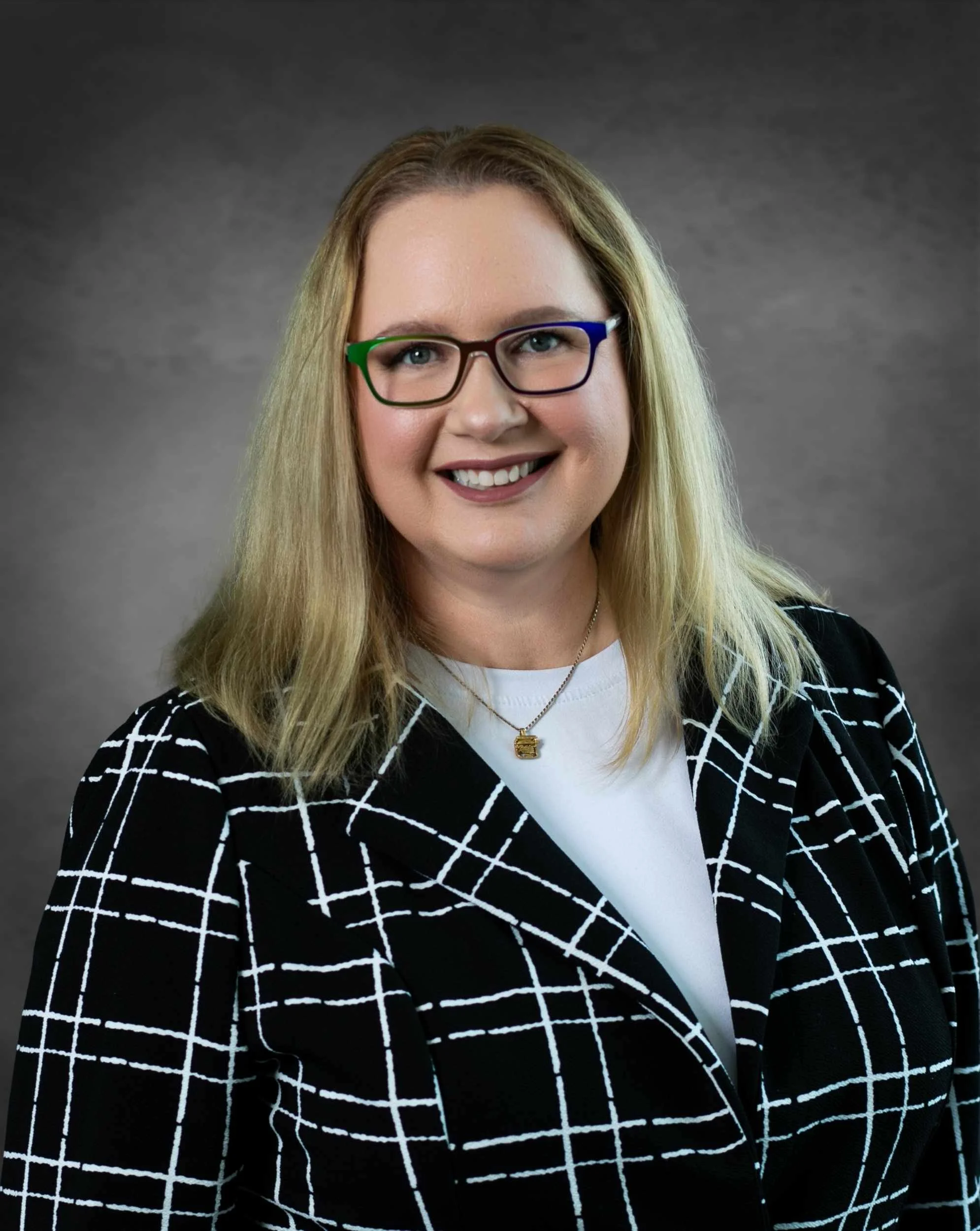 A woman with blonde hair wearing glasses with black, green, and blue frames, smiling, dressed in a black and white checkered blazer over a white top, and wearing a gold necklace with a small pendant, against a gray gradient background.