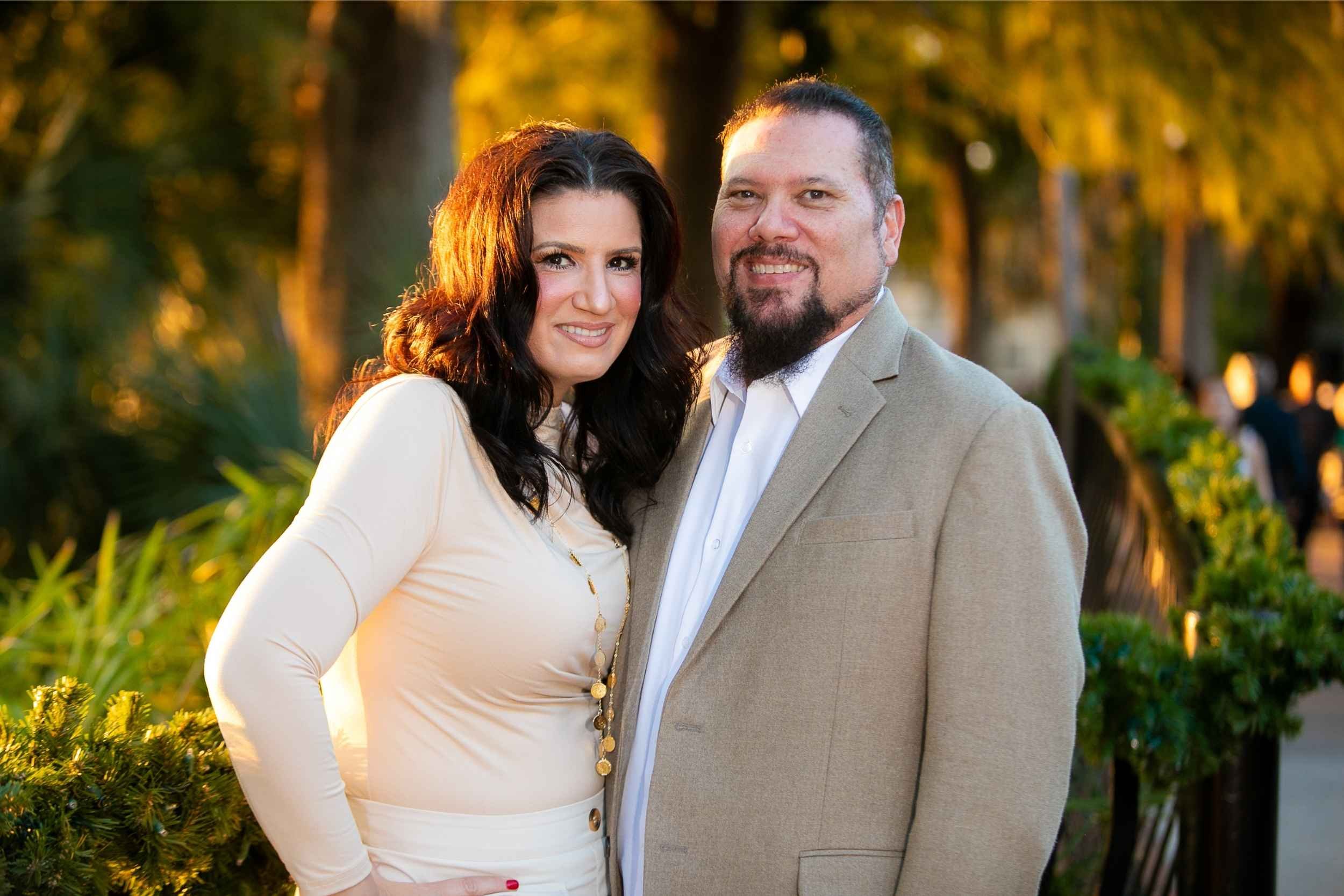 A smiling couple standing close together outdoors during sunset, with trees and plants in the background in downtown Orlando.
