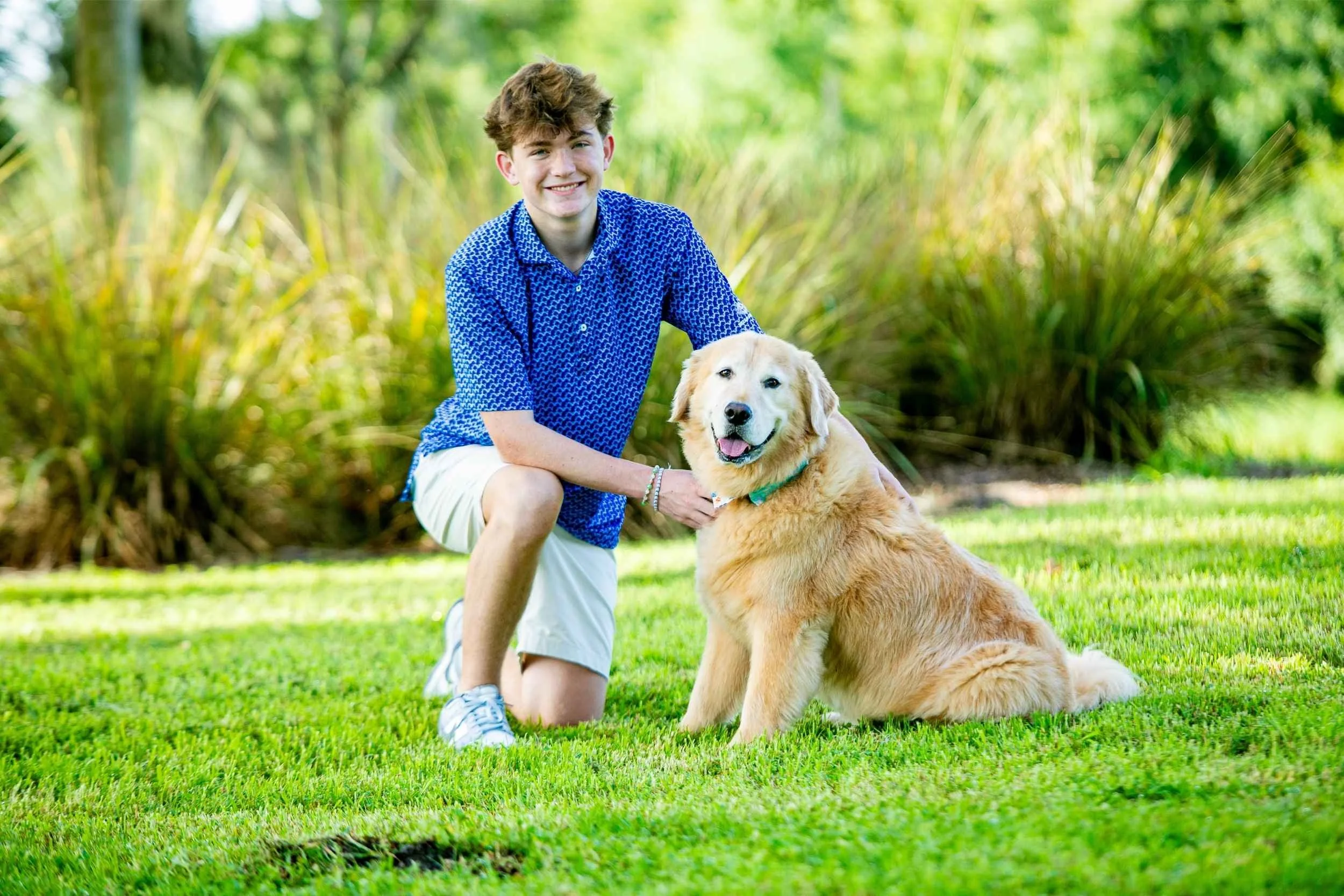 A young man kneeling with a golden retriever dog outdoor in a green park for a senior portrait session with Cannonfire Photography.