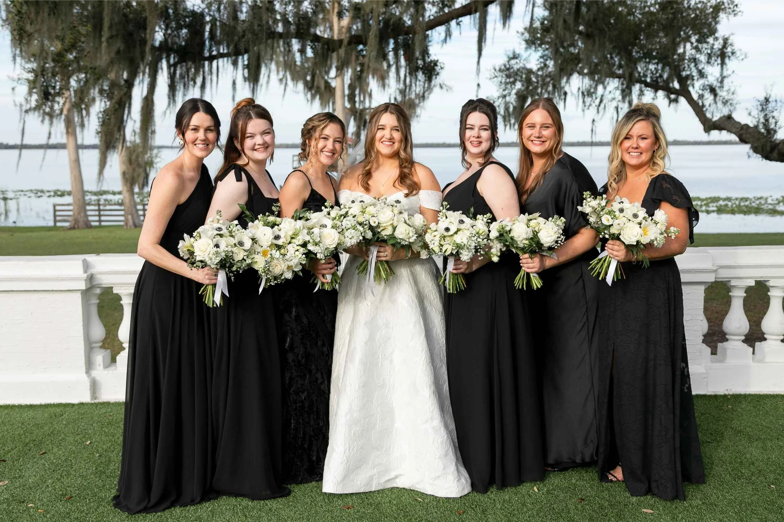Group of women in black dresses and white dress, holding bouquets of white flowers, standing outdoors near a white railing, with trees and water in the background for a wedding photographed by Cannonfire Photography.