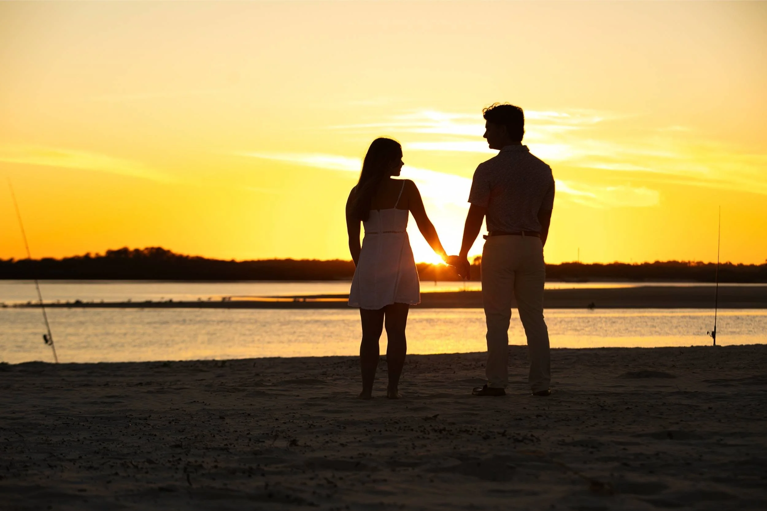 A couple holding hands on the beach at sunset, facing each other with the sun setting behind them, casting their silhouettes against the bright sky.