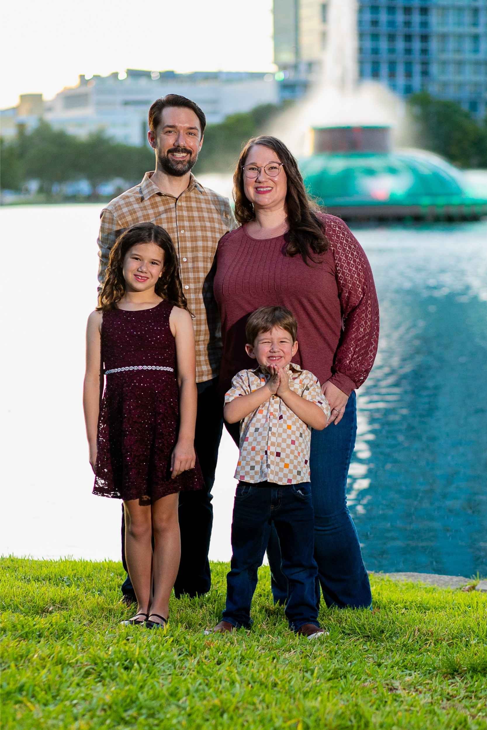 Family of four standing outdoors near Lake Eola with a fountain and downtown Orlando buildings in the background, smiling at the camera during a family portrait session with Cannonfire Photography.