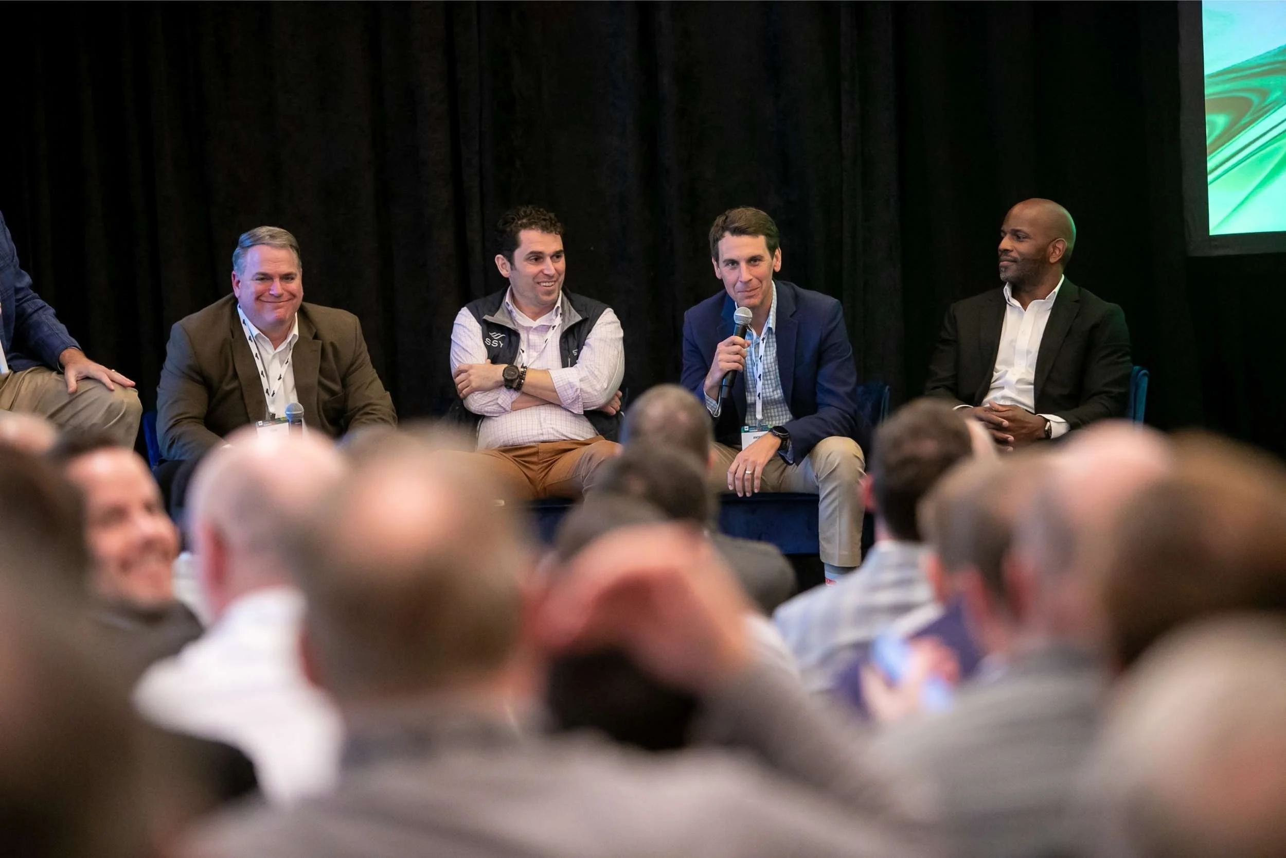 Panel of five men sitting on stage at a conference, with one speaking into a microphone and others listening, audience in foreground for a conference photographed by Cannonfire Photography.