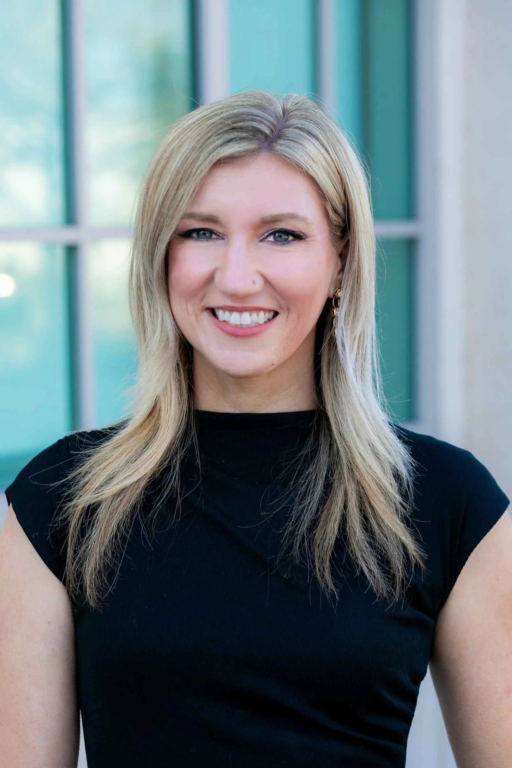 A woman with blonde hair wearing a black top, smiling in front of a green-tinted window for a headshot.