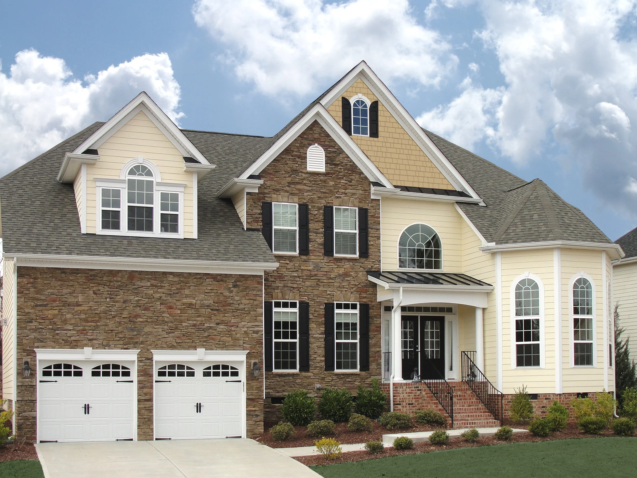 Large two-story house with a combination of stone, beige siding, and shingles exterior, multiple windows, a front porch with steps, and a two-car garage.