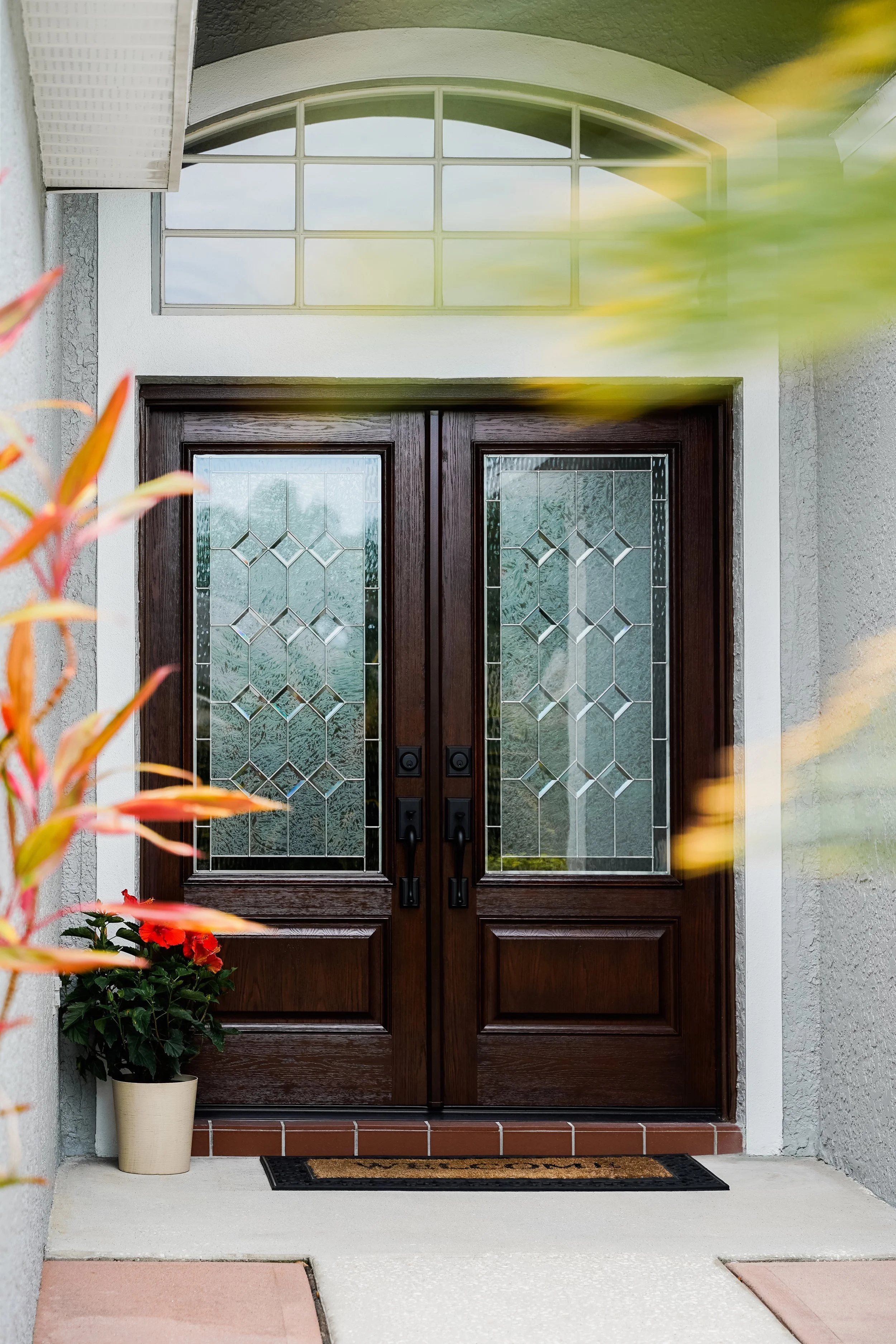 Front entrance with wooden double doors featuring glass panels, a potted red flower plant on a mat, and an arched window above.