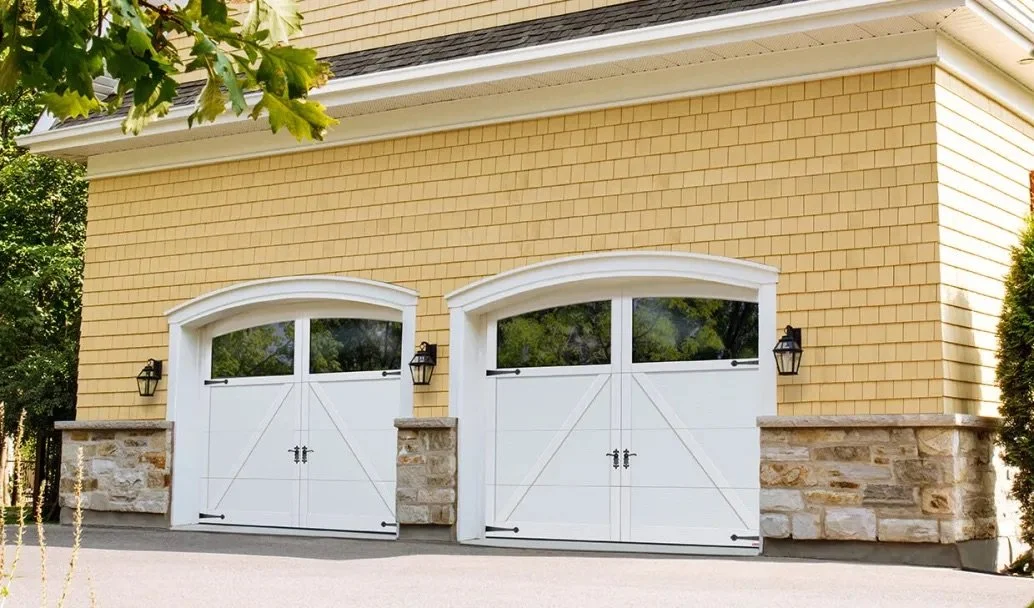 Two white garage doors with black hinges and locks, located on a yellow house with stone accents and two black wall-mounted lanterns on either side.