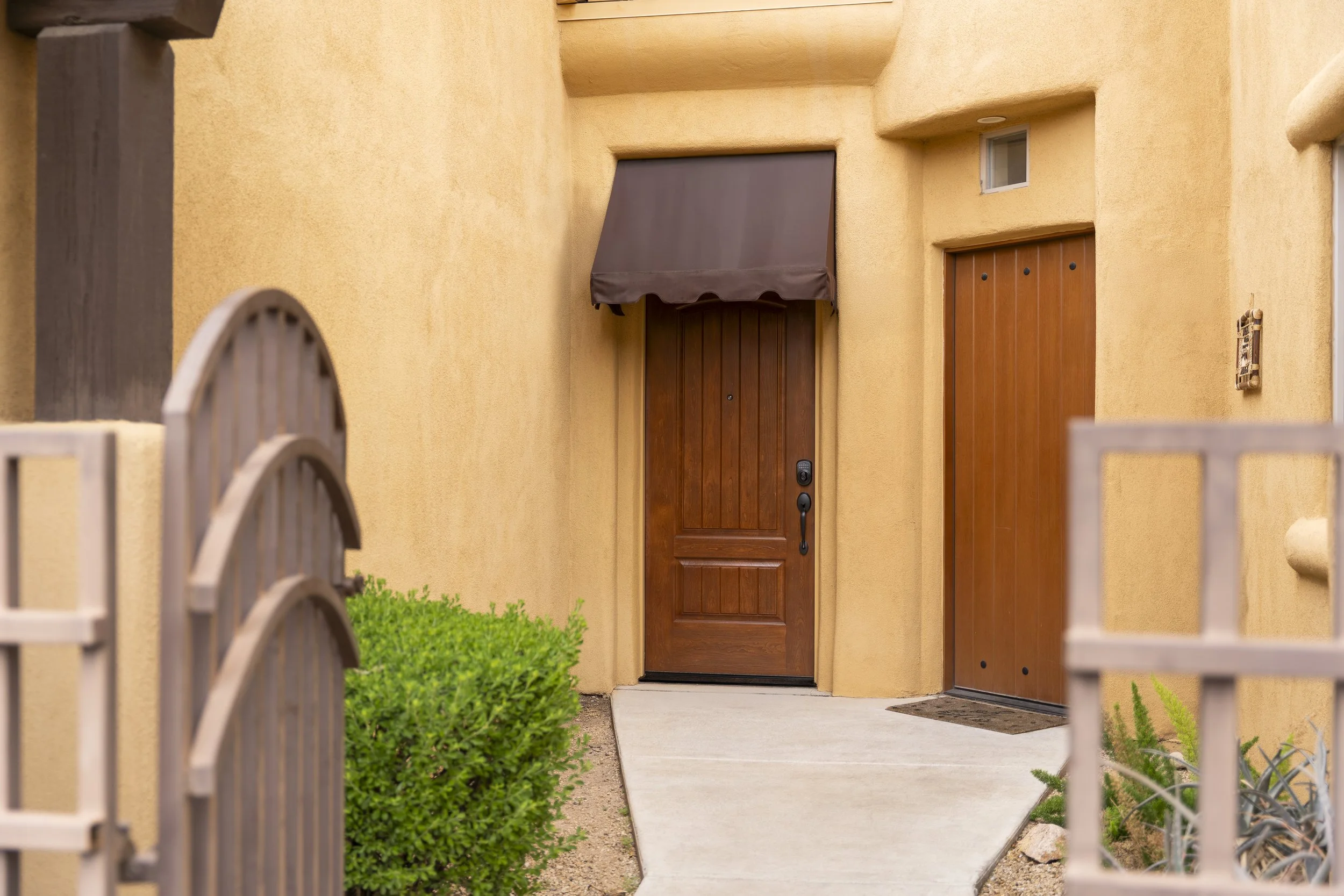 Close-up of a house entrance with a wooden front door, a dark brown awning, and a small window above. The house has a warm yellow stucco exterior. There is a concrete pathway and some greenery to the sides of the door. Part of a metal fence is visibl