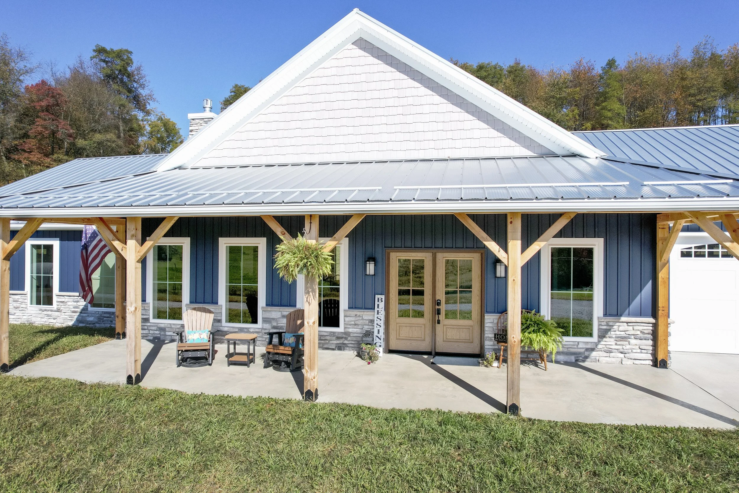 Front of a modern house with a covered porch, blue siding, stone accents, and a metal roof, decorated with outdoor furniture and potted plants.