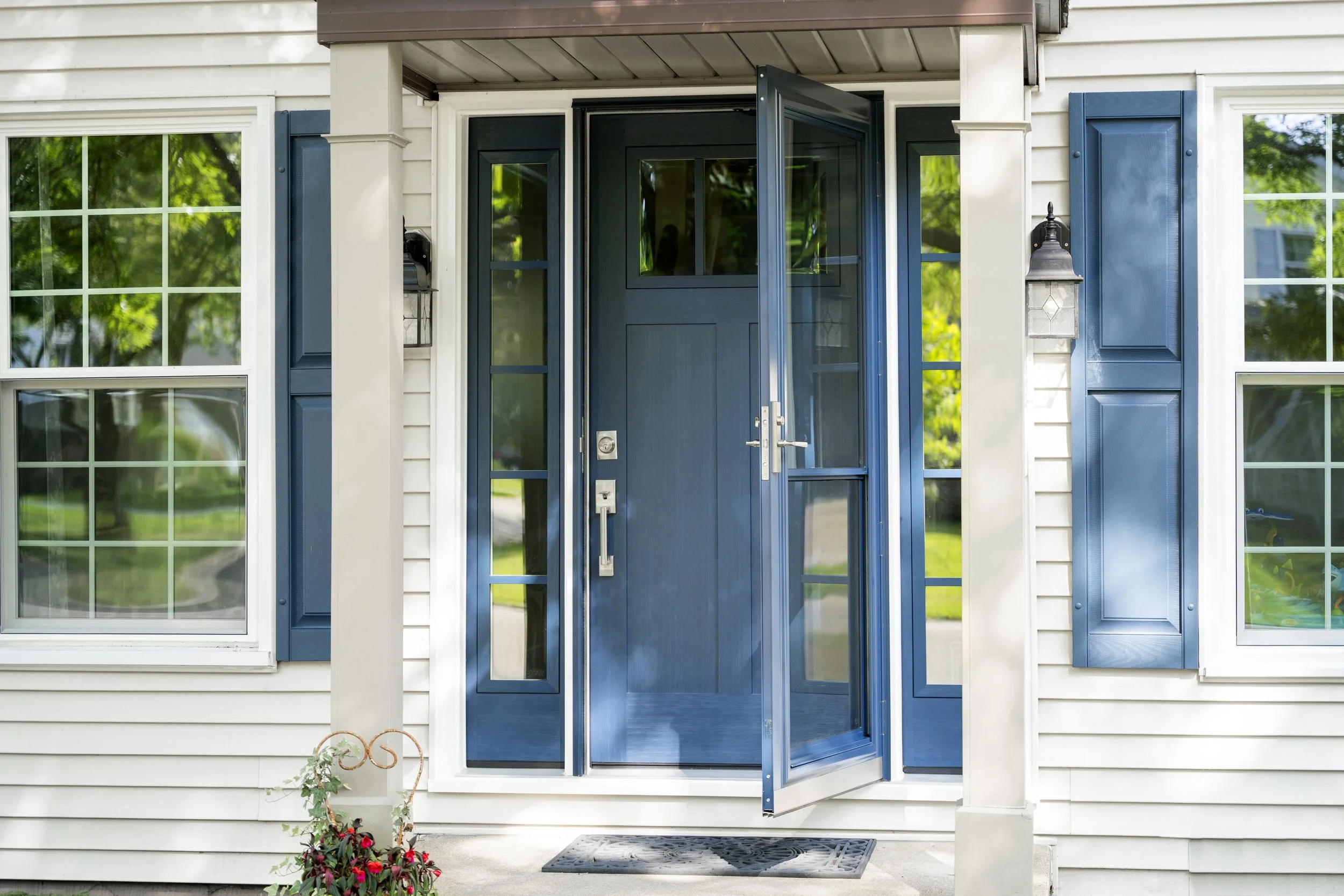 Front entrance of a house with a blue door and matching blue window shutters, flanked by two white columns and lit by a black outdoor lantern