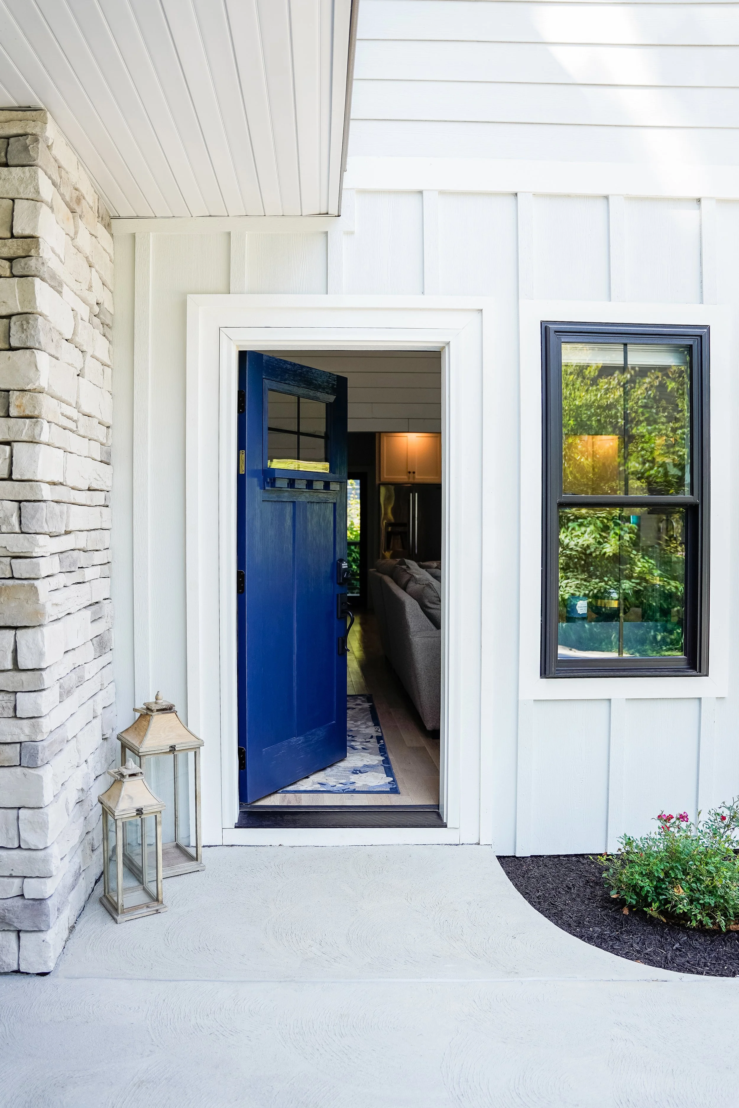 Open front door of house revealing living room with couch and kitchen in background, blue door with window, white exterior walls, window with black trim, and decorative lanterns outside.