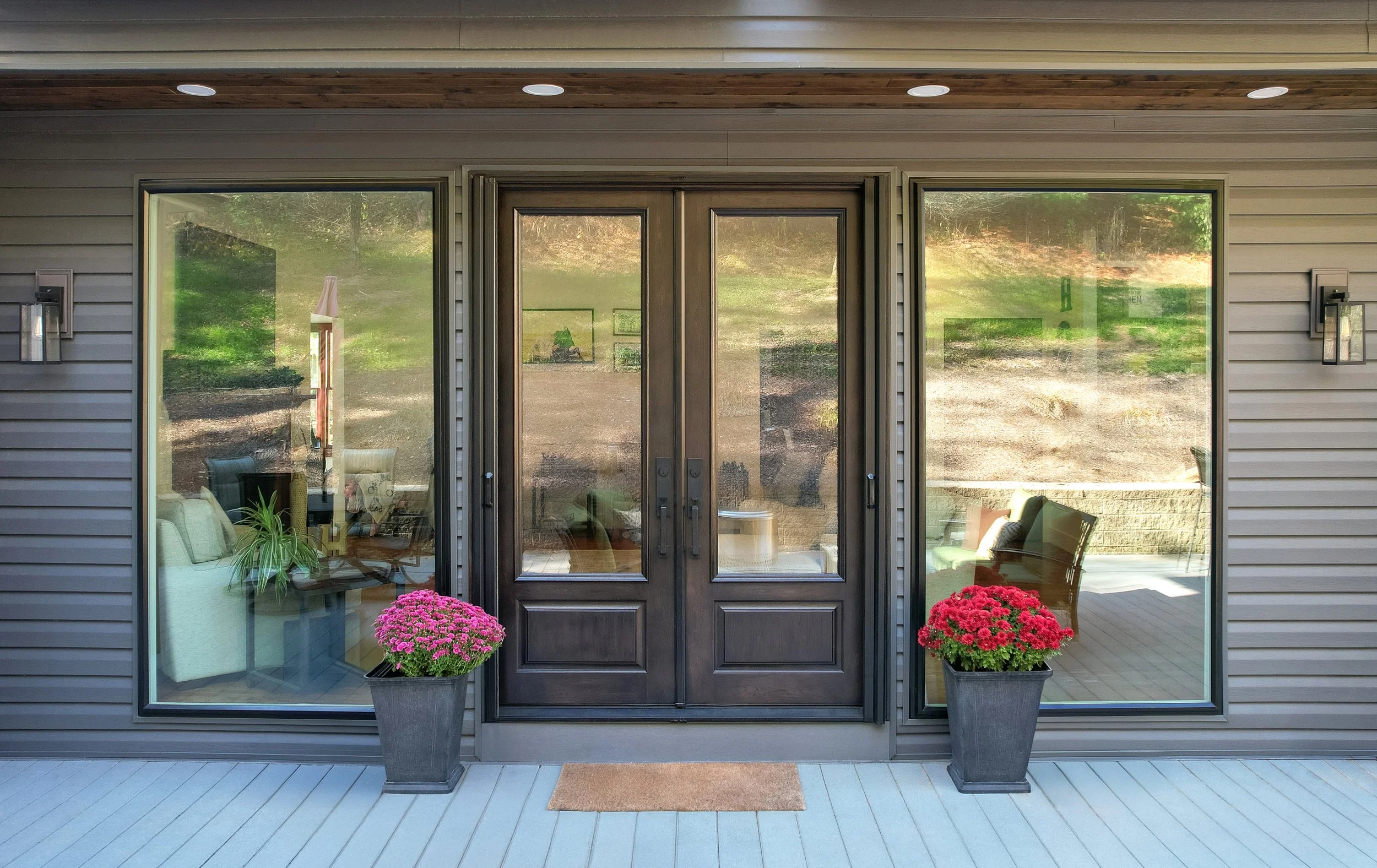 Front porch of a house with large glass windows, black double doors, two gray flower pots with pink and red chrysanthemums, a brown doormat, and beige siding.