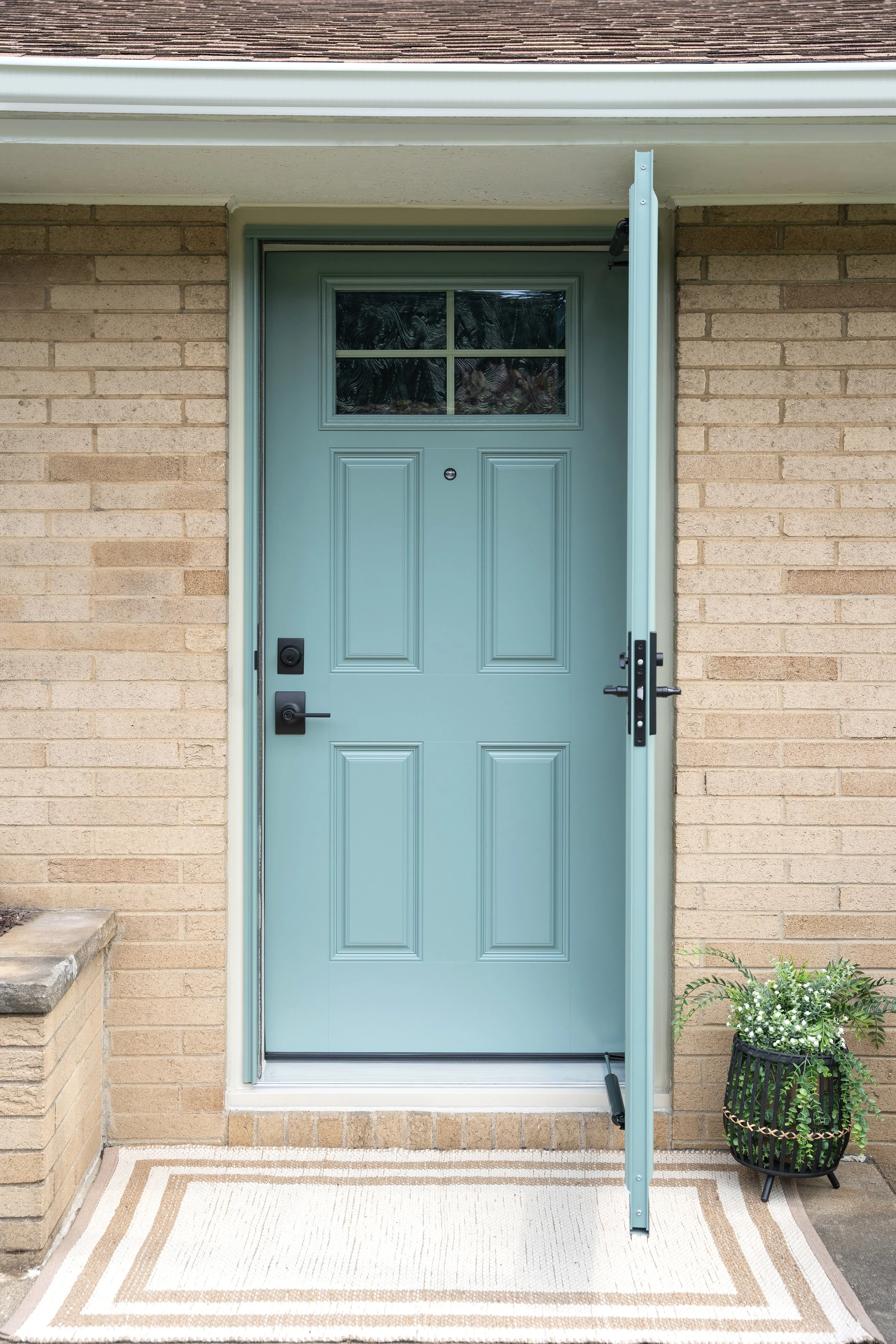 Blue front door with window, open storm door, brick house exterior, potted plant with white flowers on right, beige doormat with geometric border pattern.