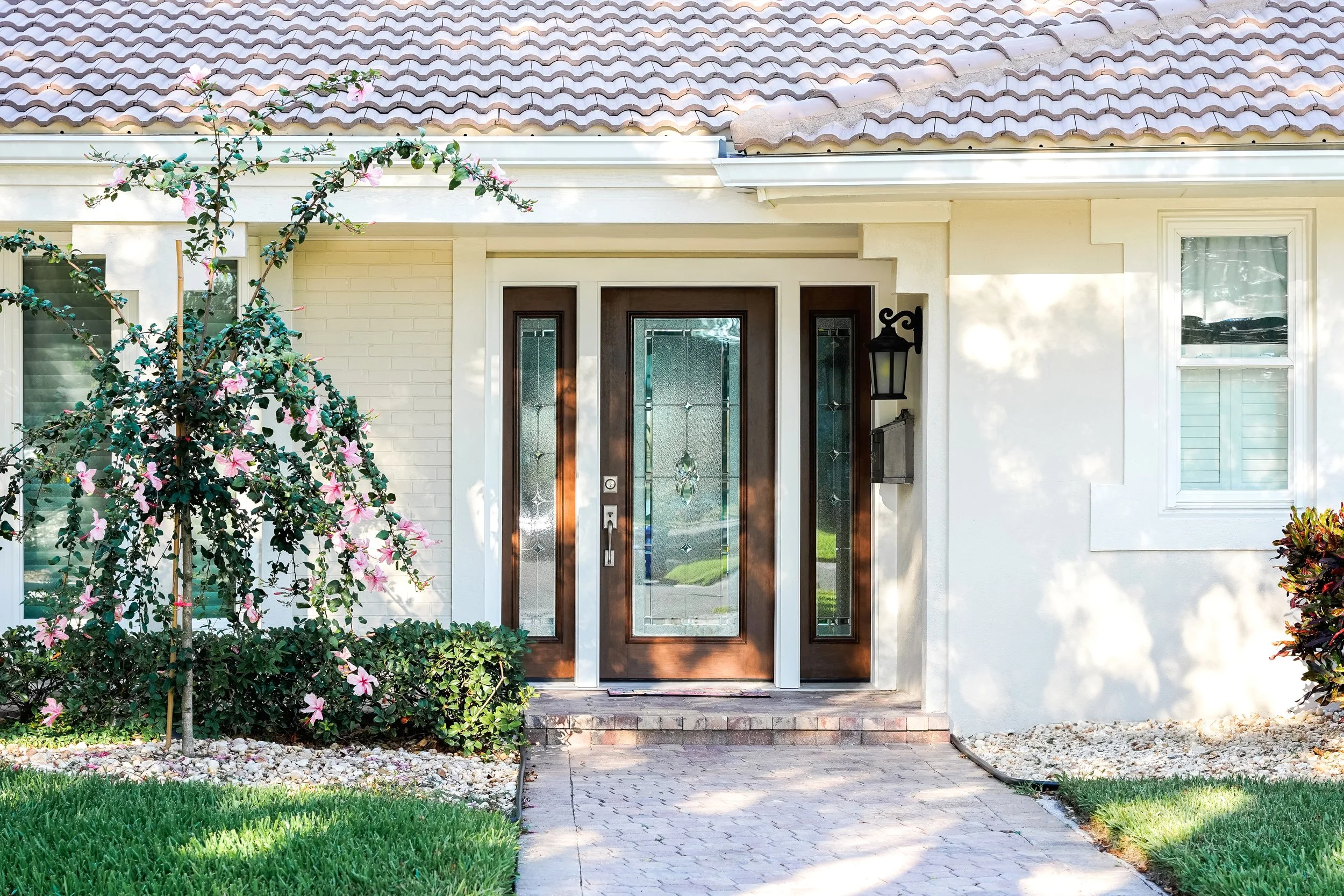Front door of a house with decorative glass panels, surrounded by greenery, flowering plant, and a brick walkway.